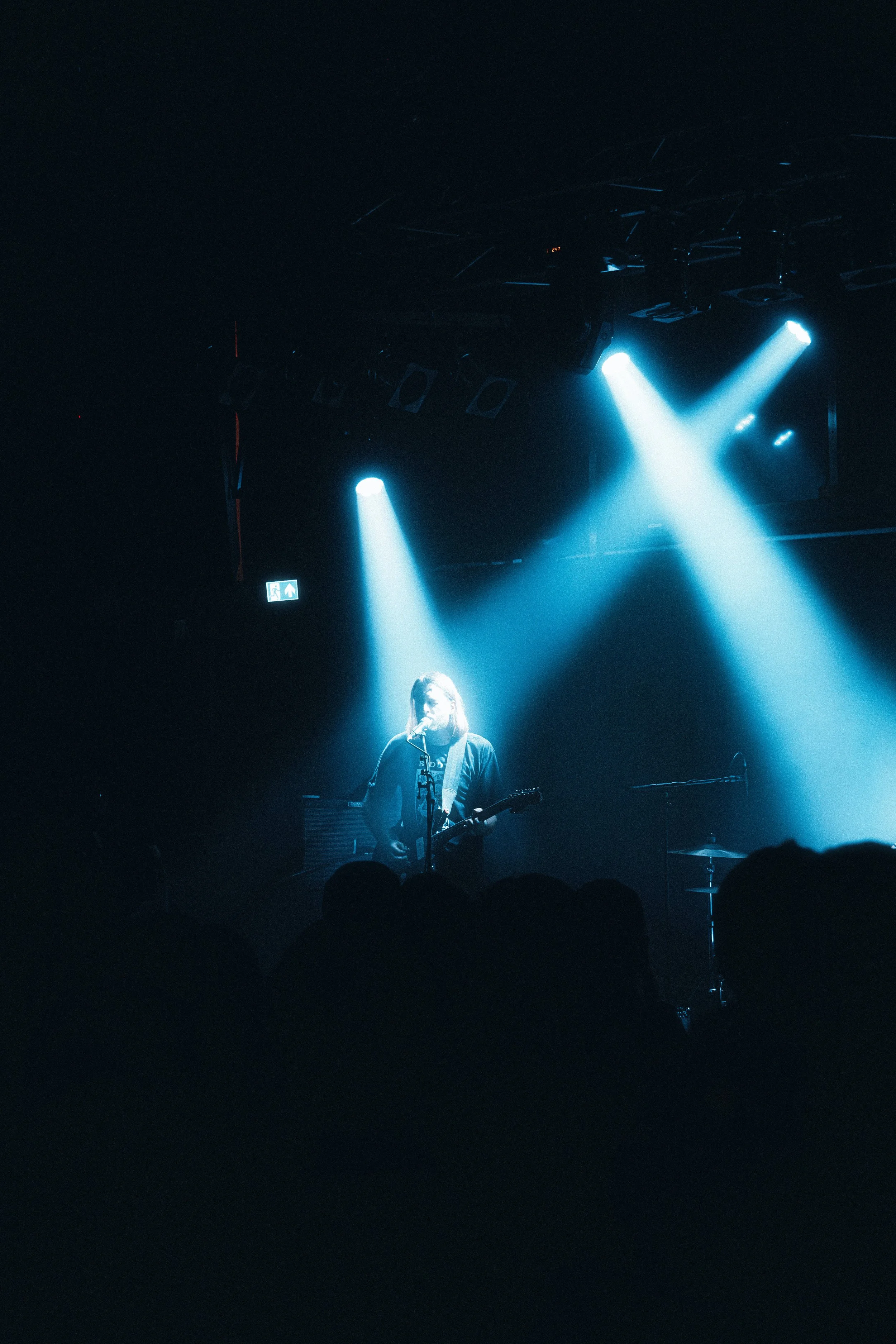 A musician performing on stage with a guitar under beams of blue stage lighting in front of a dark audience.