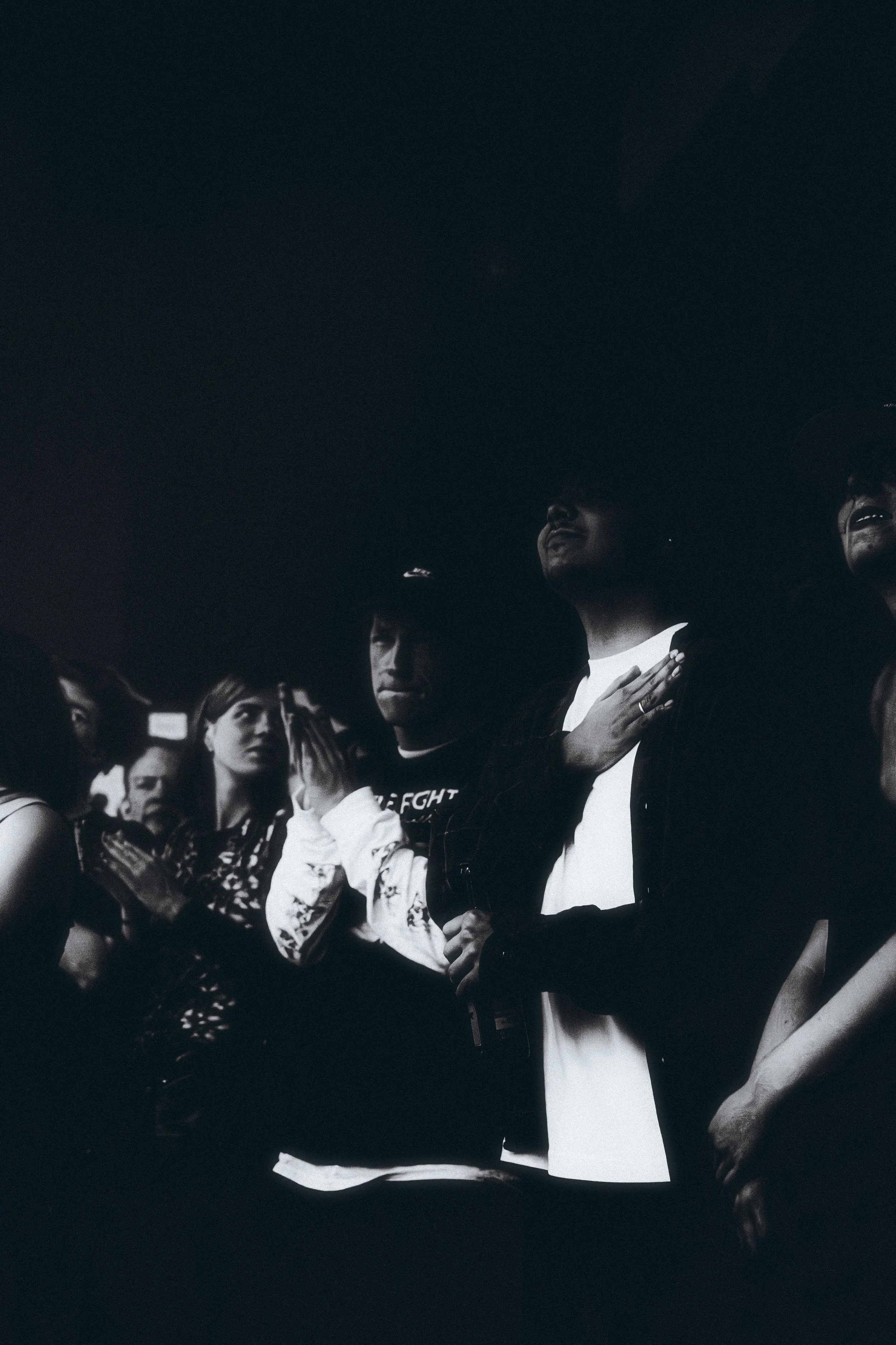 A group of people standing closely together with their hands on their hearts, looking upward, in a dark setting.
