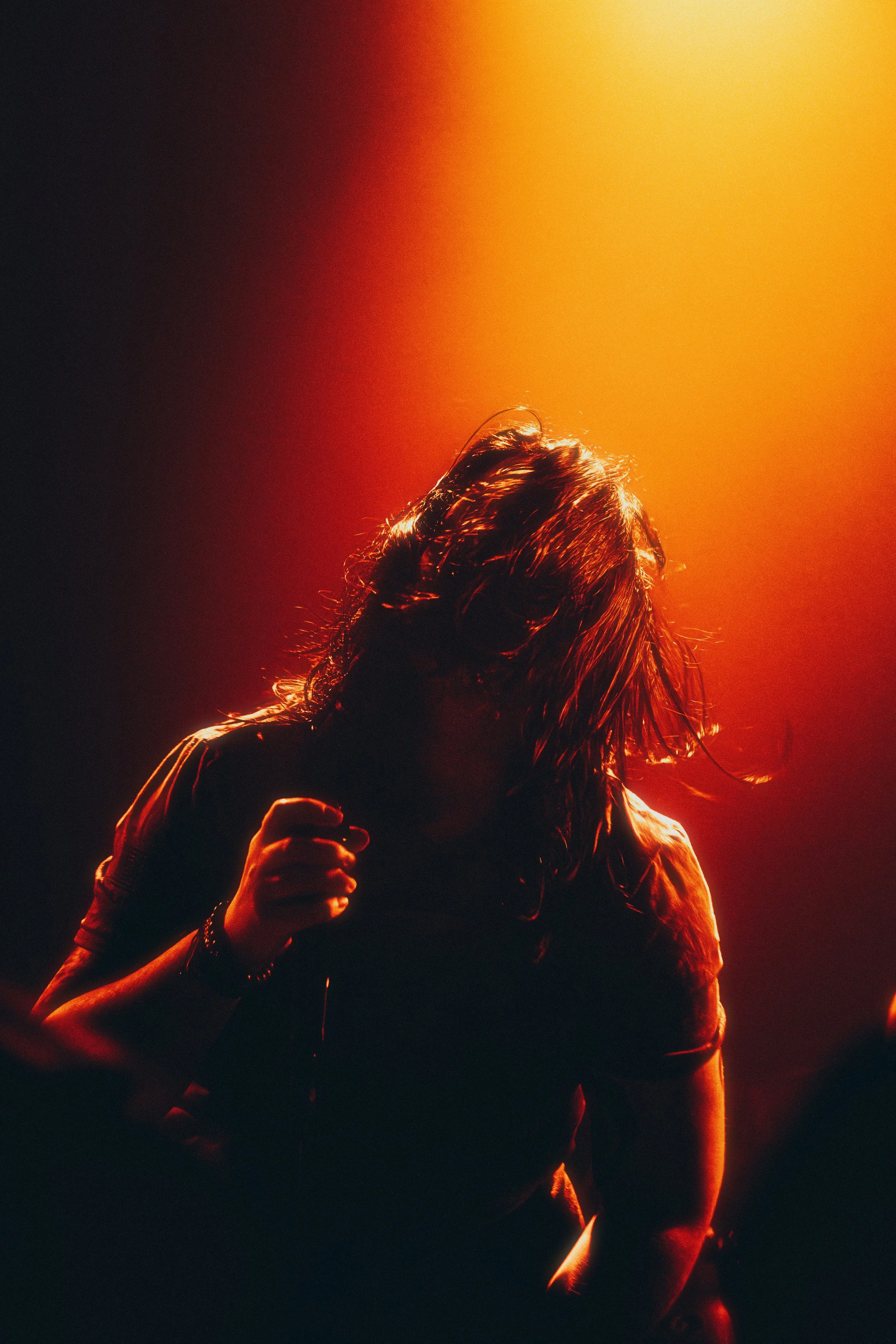 A person with long hair dancing or performing on stage, illuminated by warm orange and yellow stage lighting, with their head down and hair covering their face.