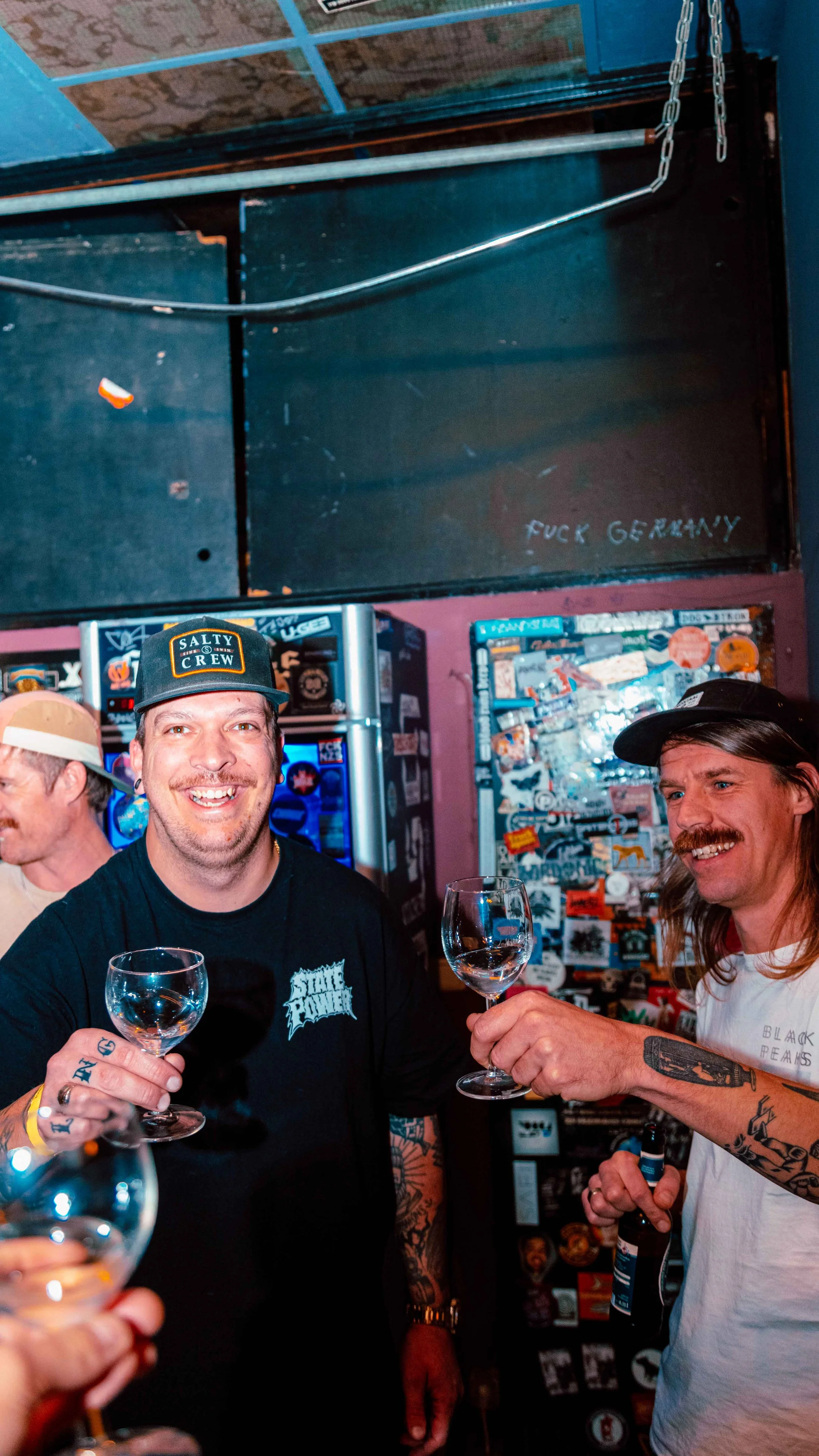 Two men smiling and toasting with glasses of wine or spirits in a bar, with stickers on the fridge behind them and a dark wall above with graffiti that reads 'FUCK GERMANY'.