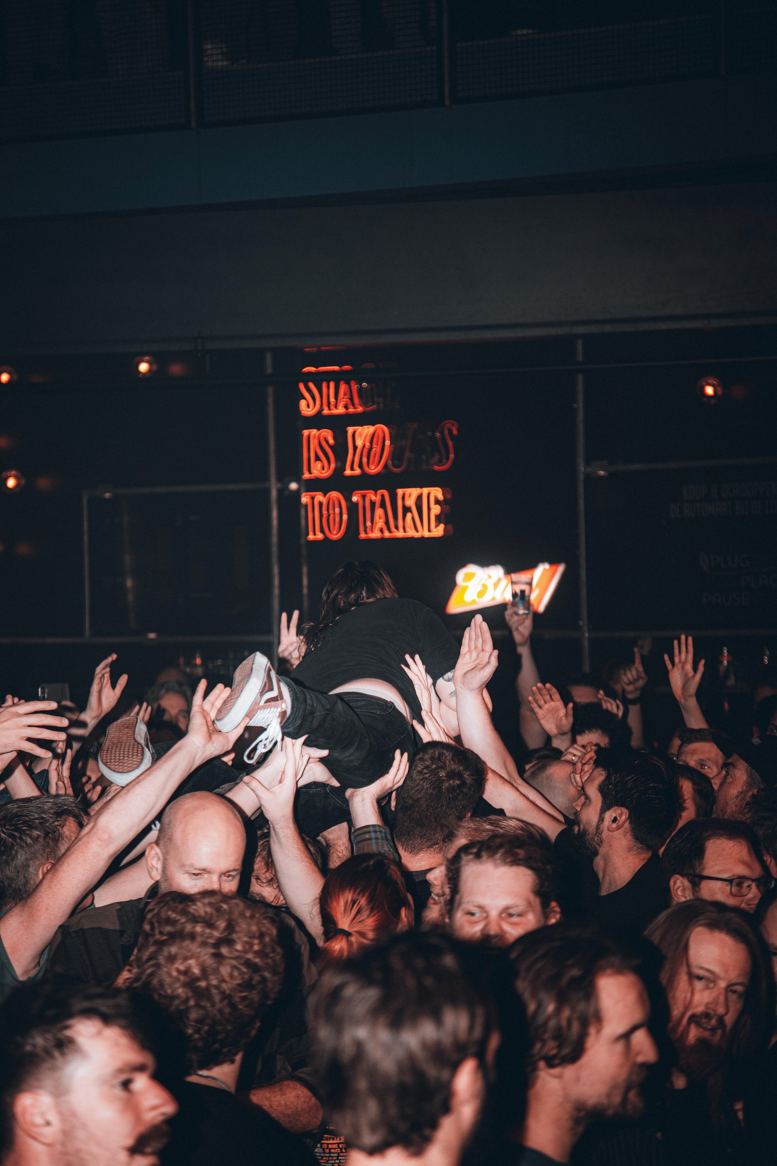 Crowd of people at a concert or event, with one person being crowd-surfed above the others. Neon sign in the background reading 'Silence is not always to take'.
