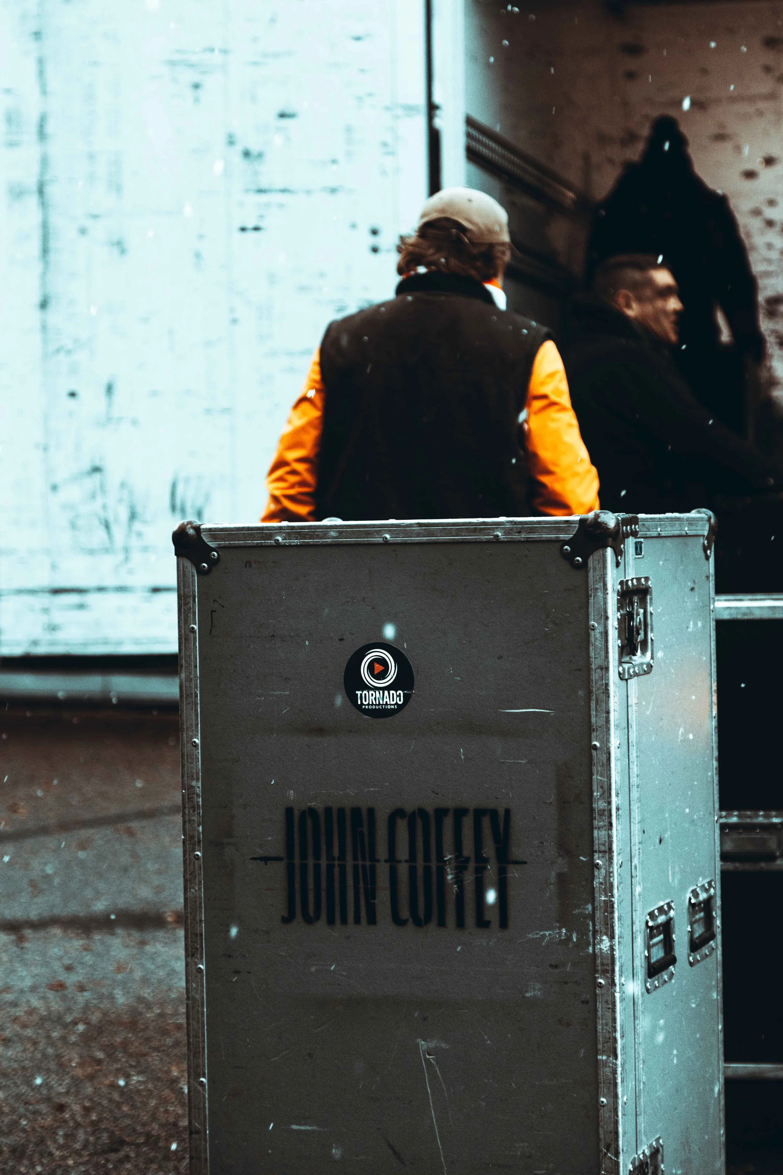 Outdoor scene with two men, one in an orange and black jacket and gray cap, standing behind a large equipment case with John Cofty name and Tornado Productions logo. The background features a distressed wall and some equipment.
