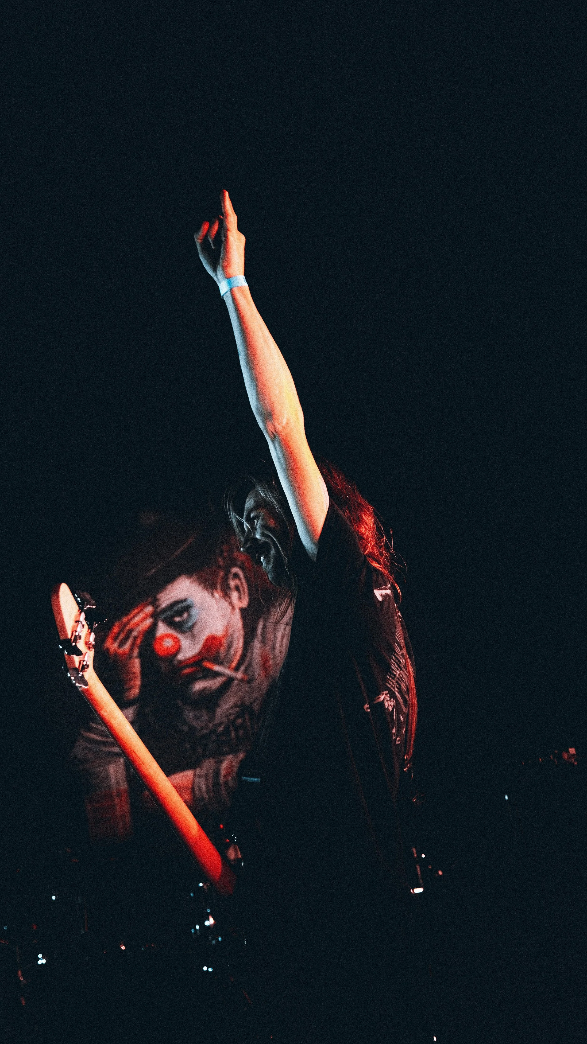 A man with clown makeup and long hair playing an electric guitar with one arm raised in the air during a dark concert.