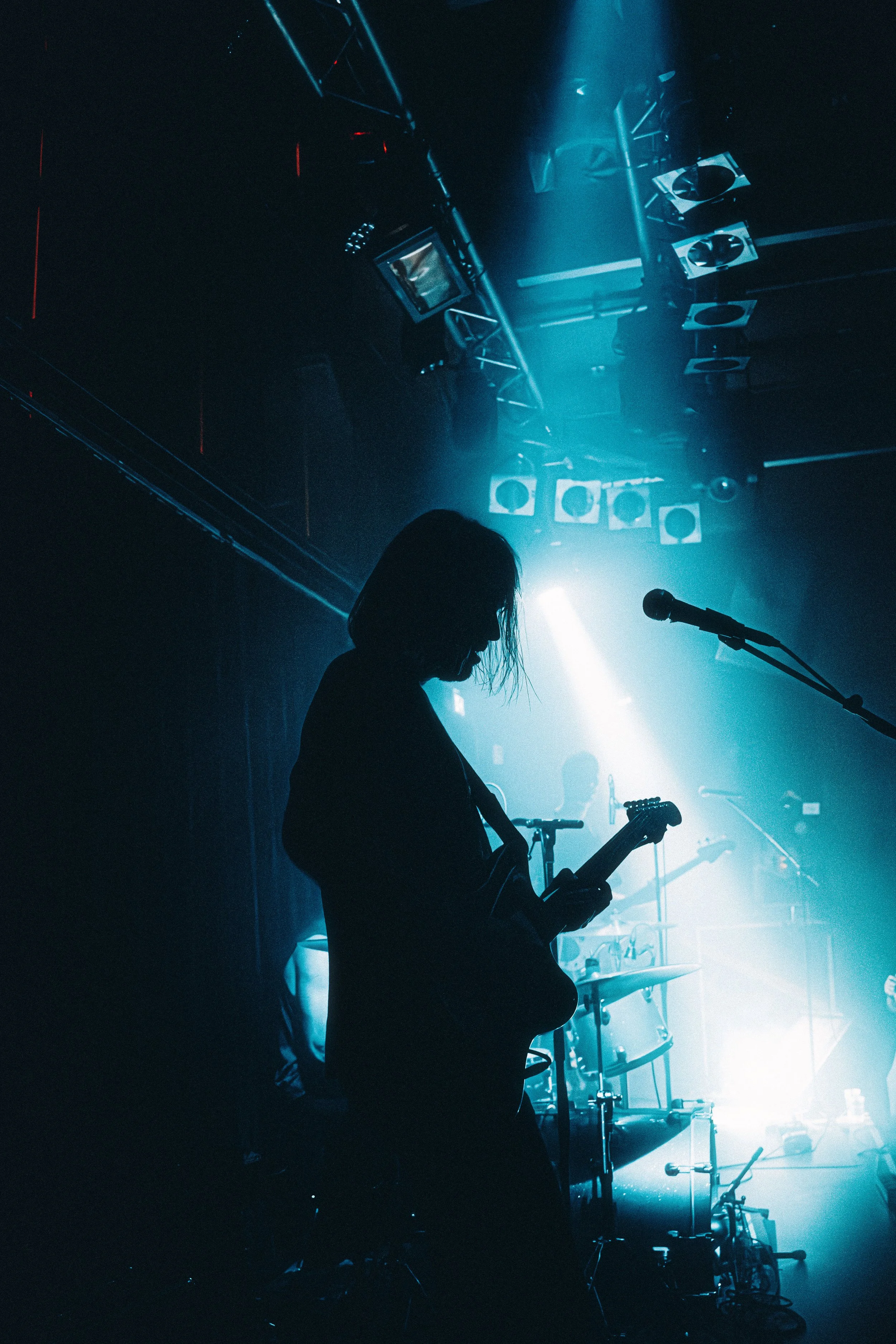 Silhouette of a female musician playing an electric guitar on stage during a live concert, with band members and stage lights in the background.