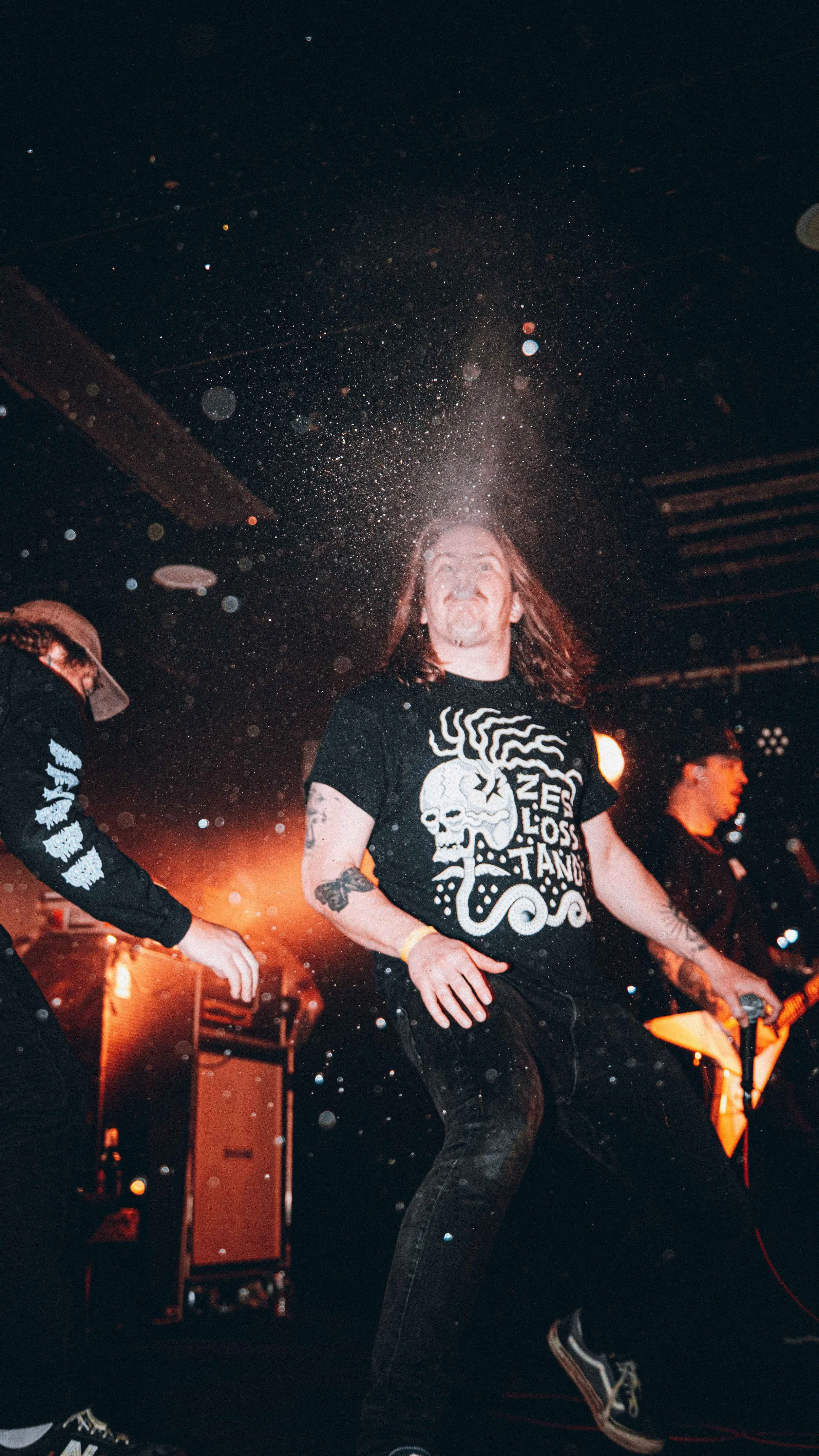 A band performs live on stage with one member in the foreground smiling, wearing a Black T-shirt with a skull and octopus graphic, and other band members playing guitars in the background, with stage lights and equipment visible.