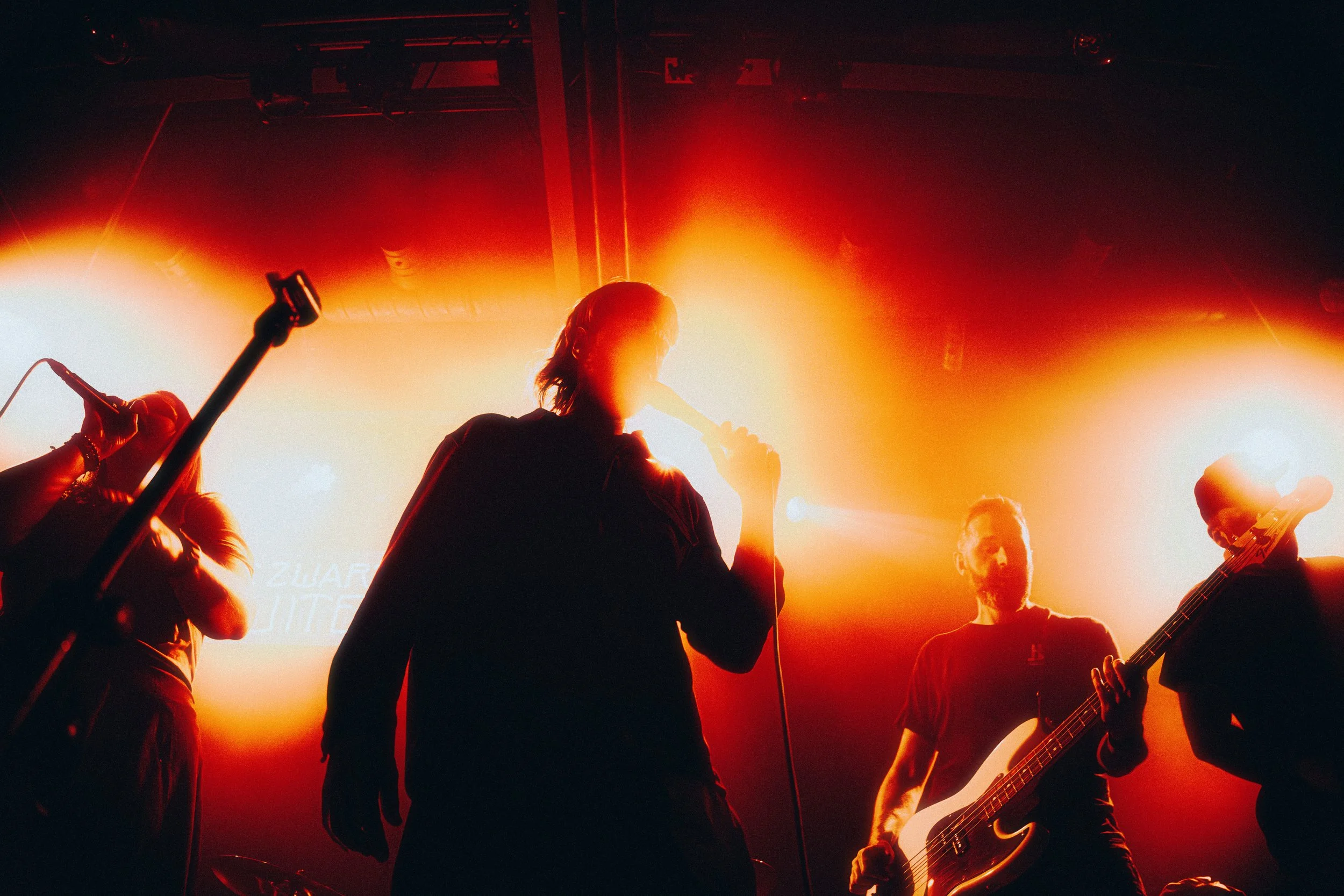 Musicians performing on stage with bright orange and red lighting, one holding a microphone, another playing bass guitar, with silhouettes and stage equipment visible.