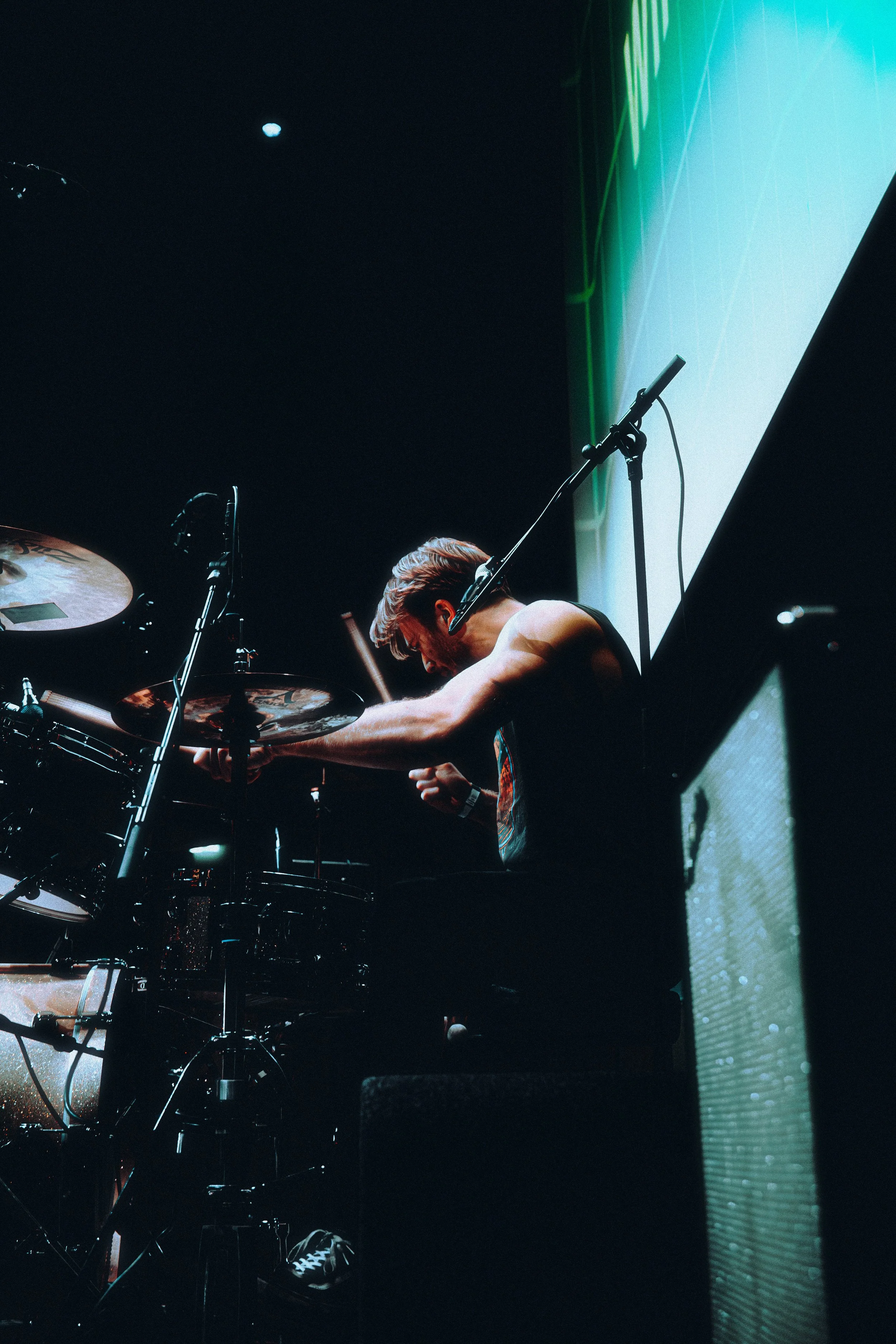 A musician playing drums on stage, bent over with headphones, illuminated by stage lighting, with a large screen in the background.