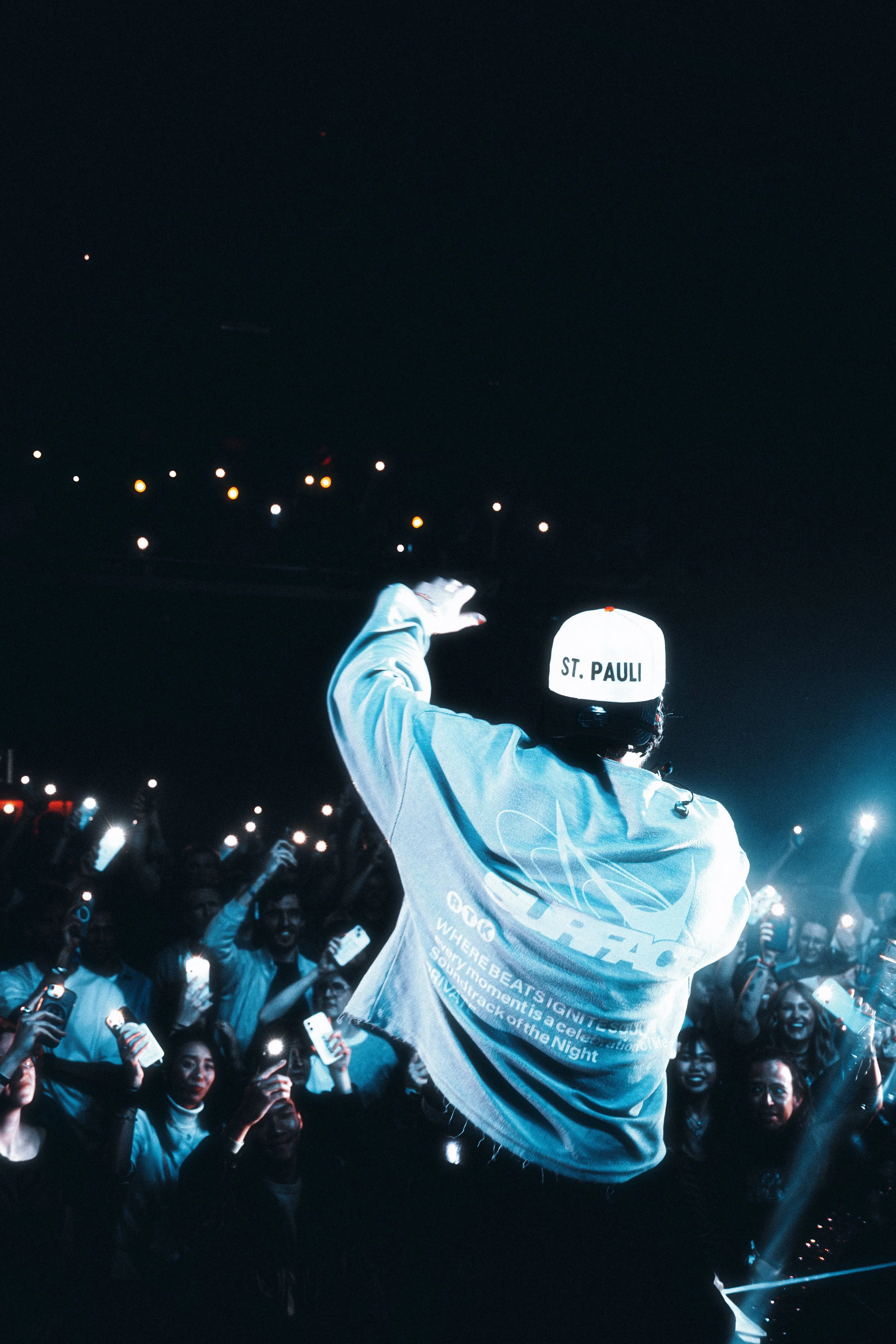 A performer on stage with back to camera, wearing a white cap with 'ST. PAULI' and a gray jacket, facing a crowd at a concert or event.
