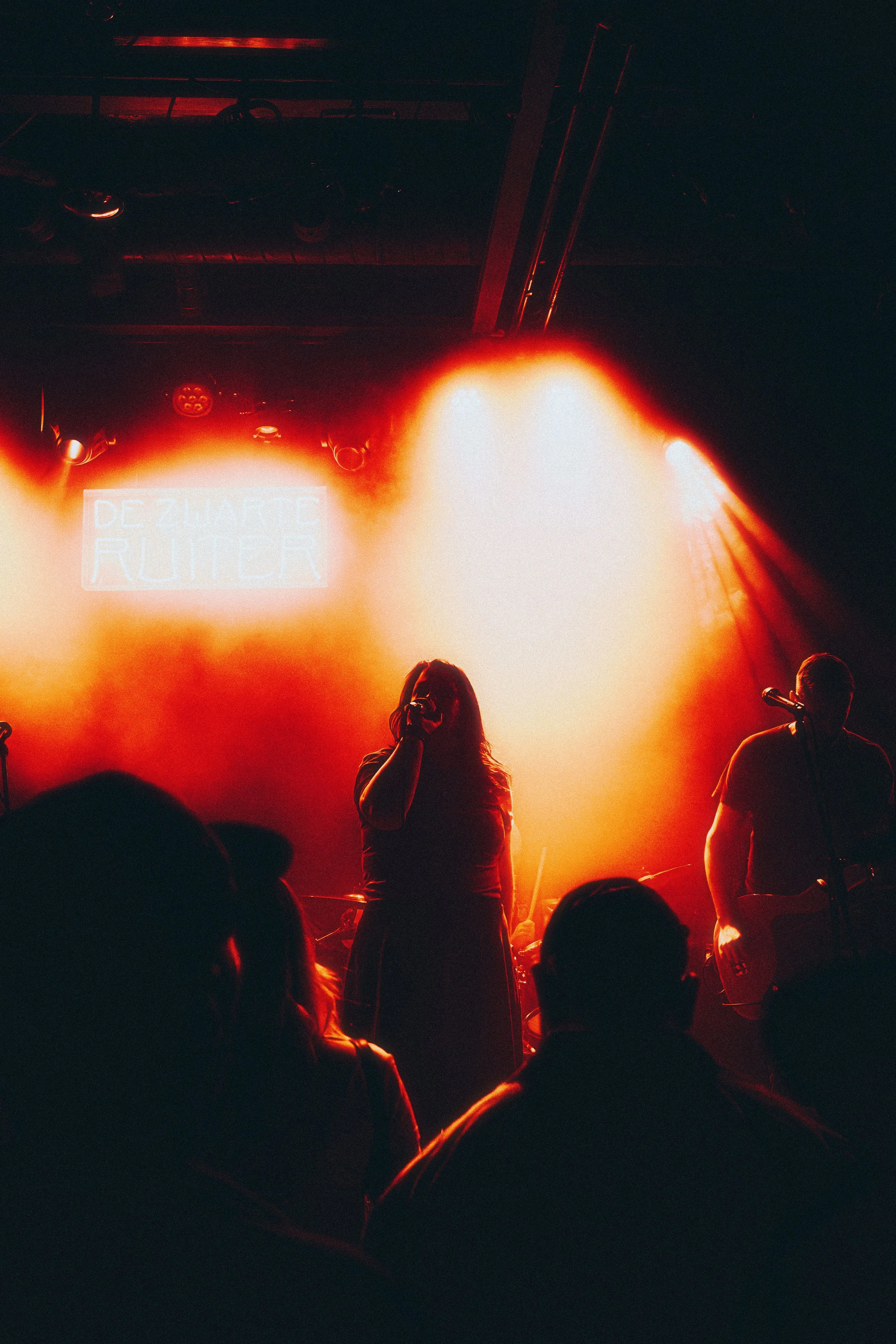Silhouette of a female singer holding a microphone on stage with bright backlighting, accompanied by a guitarist on his right, with a crowd in the foreground. Stage lights create an orange glow, and a sign in the background reads 'De Zwarte Ruiter'.