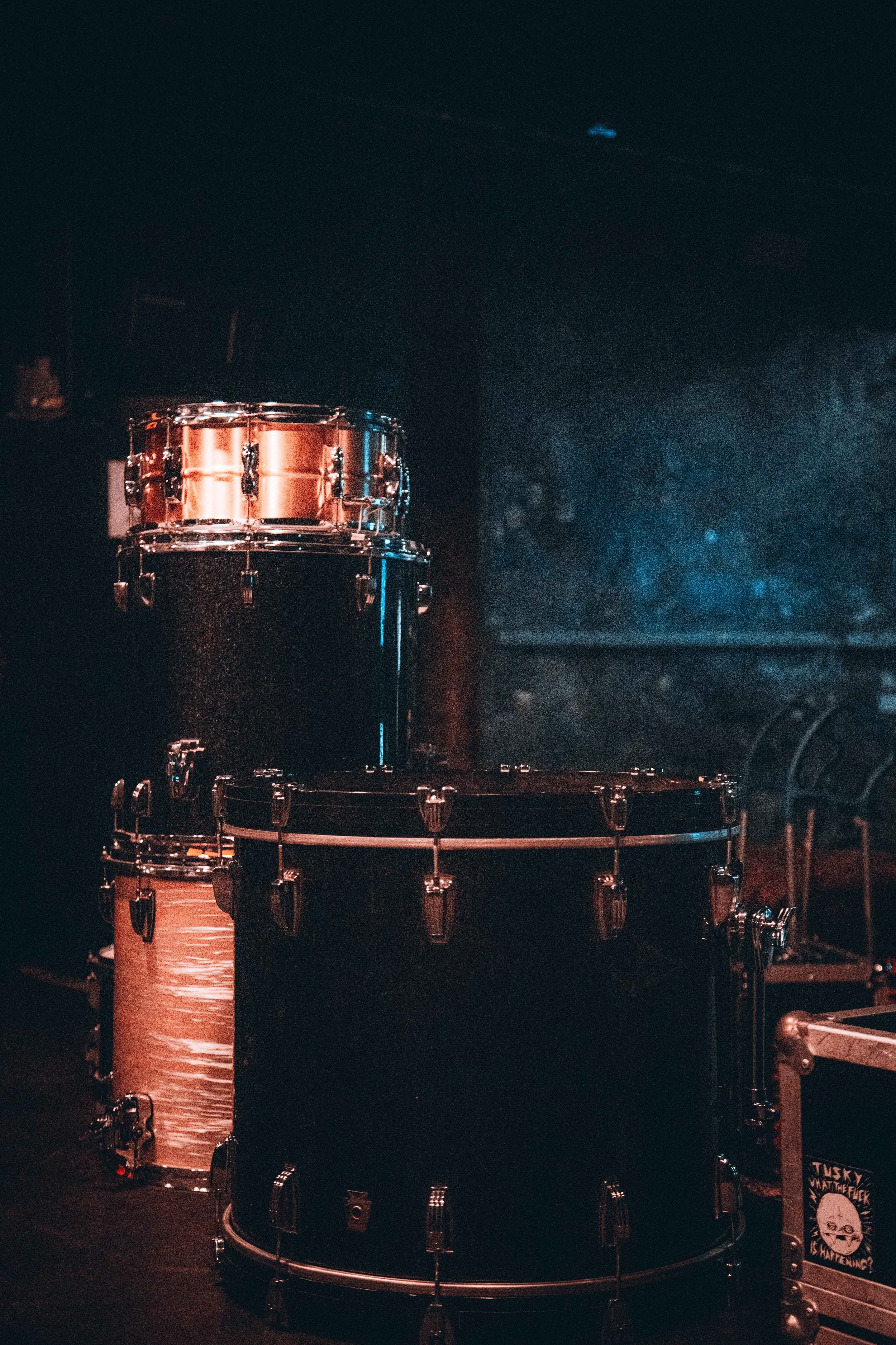 Three drums of different sizes, a small copper-colored drum at the top, a medium black drum below it, and a larger black bass drum at the bottom, on a dark stage or studio with blue lighting.