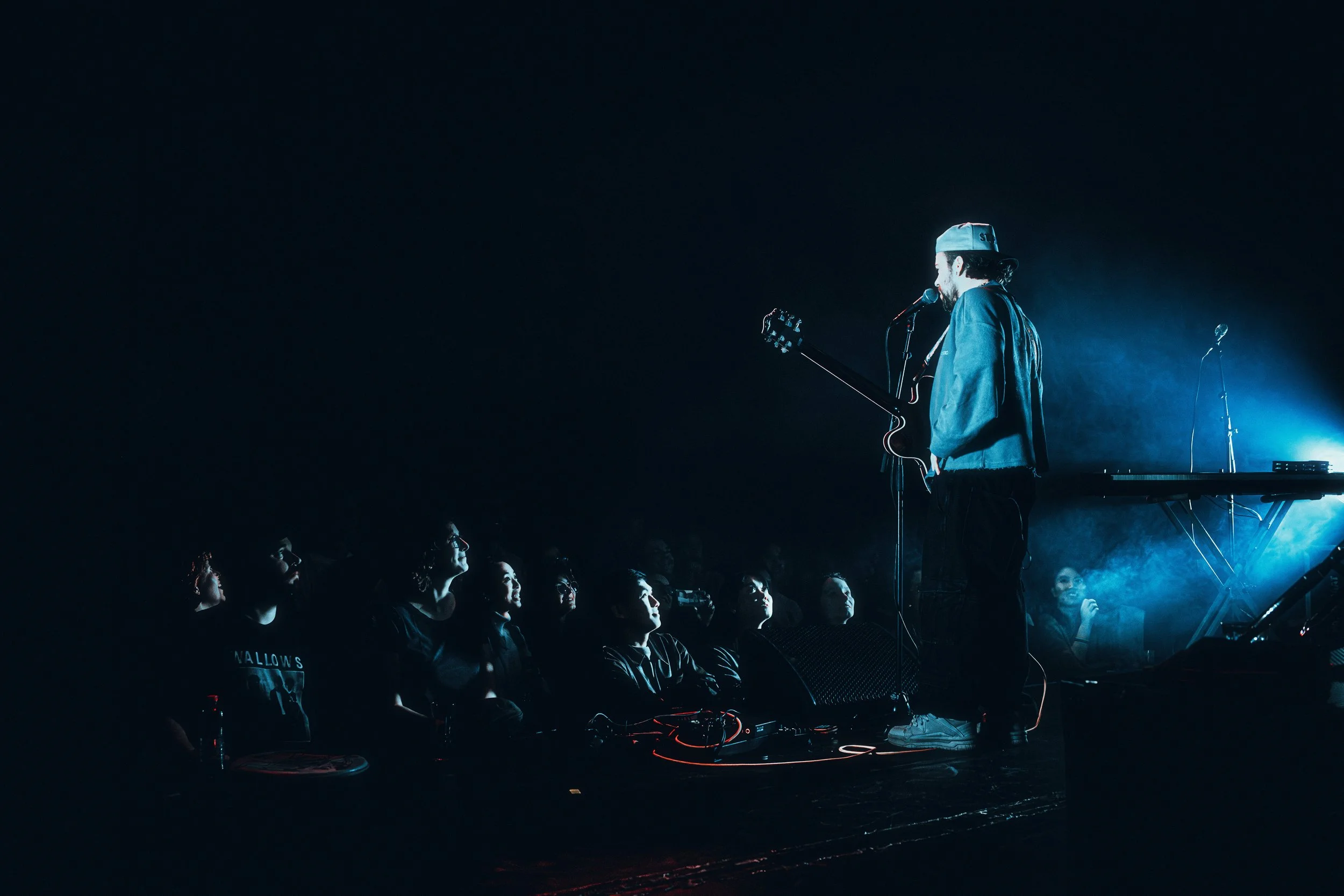 A musician stands on stage with a guitar, speaking into a microphone, illuminated by blue stage lighting, with an audience watching attentively.