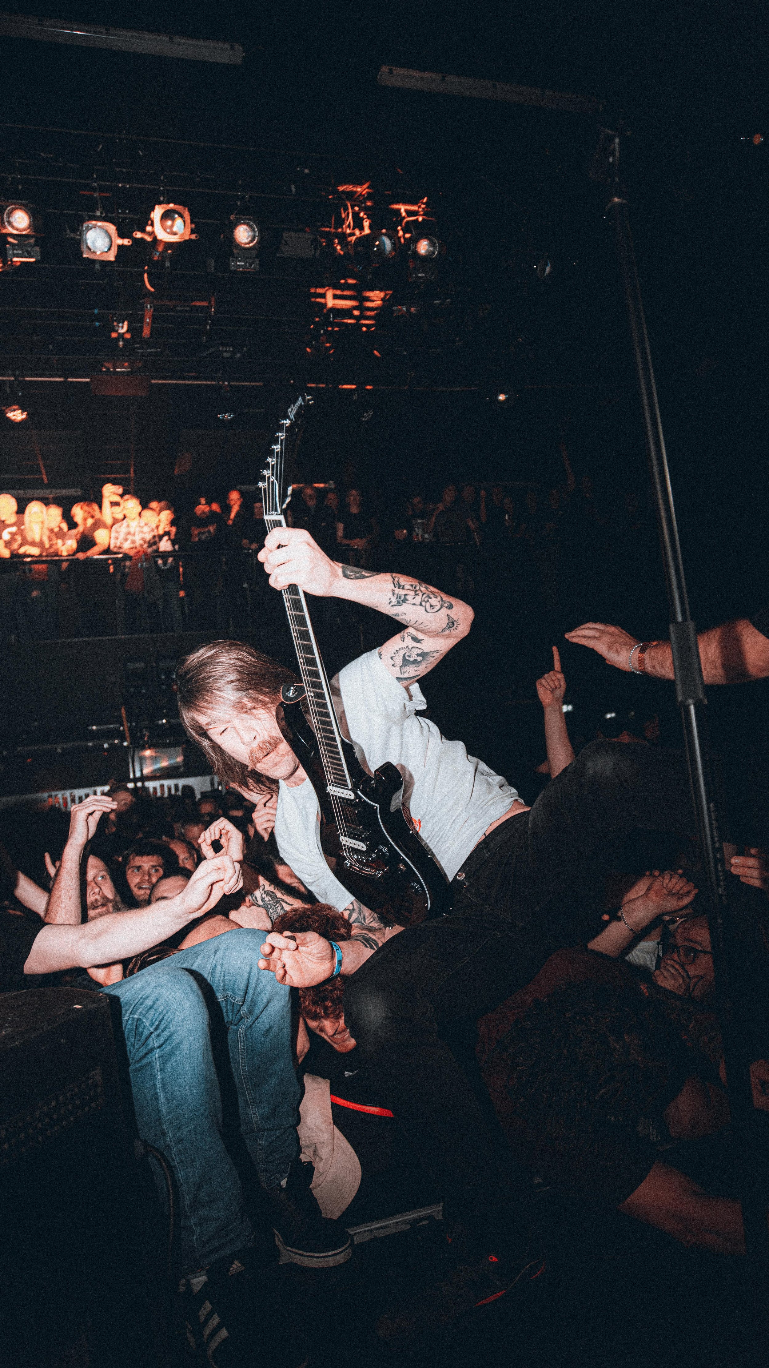Musician crowdsurfing with a black electric guitar during a live concert, audience reaching out at a crowded indoor venue.