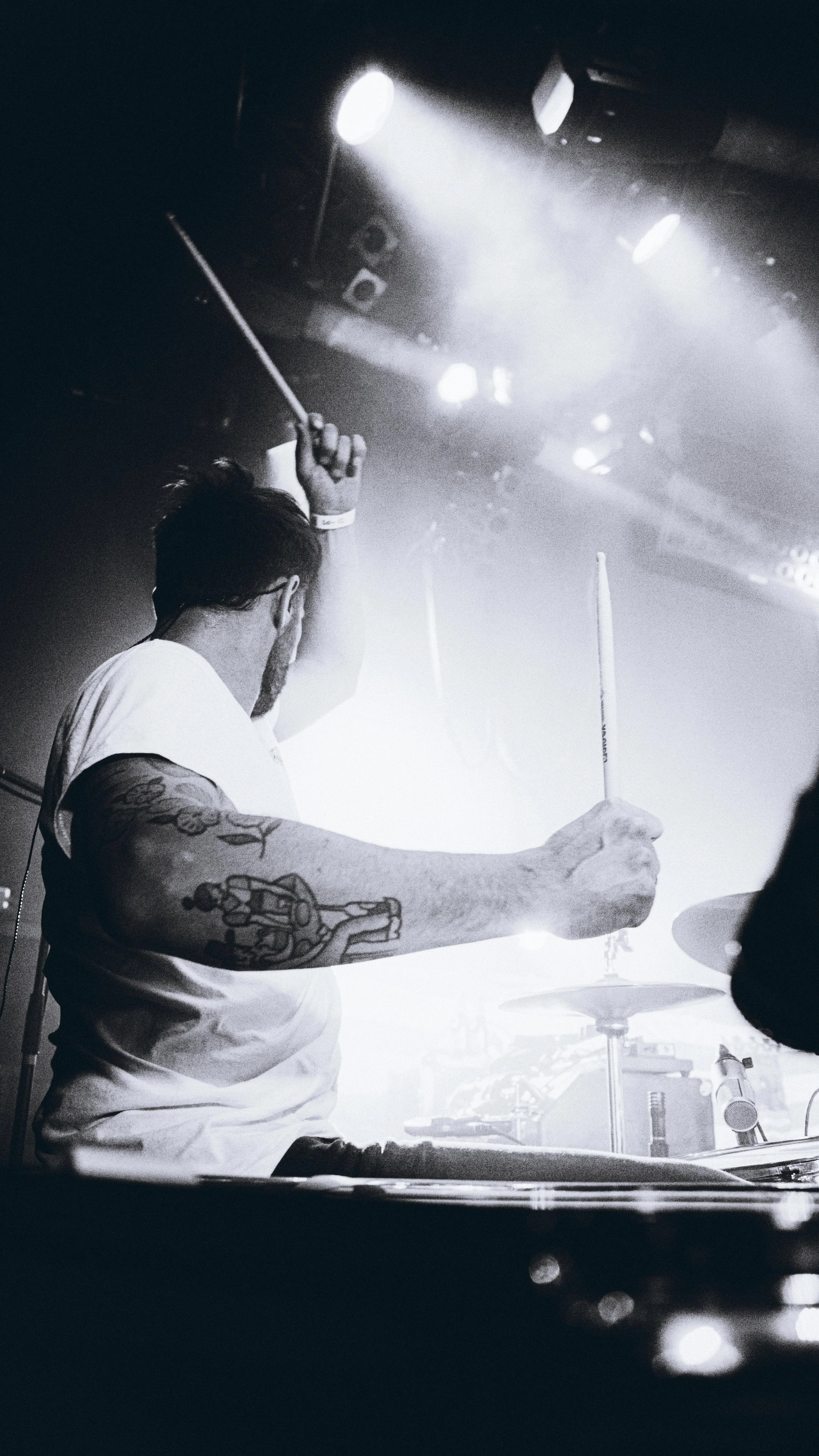 A drummer playing in a dimly lit stage, with bright lights shining down, wearing a white t-shirt and sporting tattoos on the left arm.