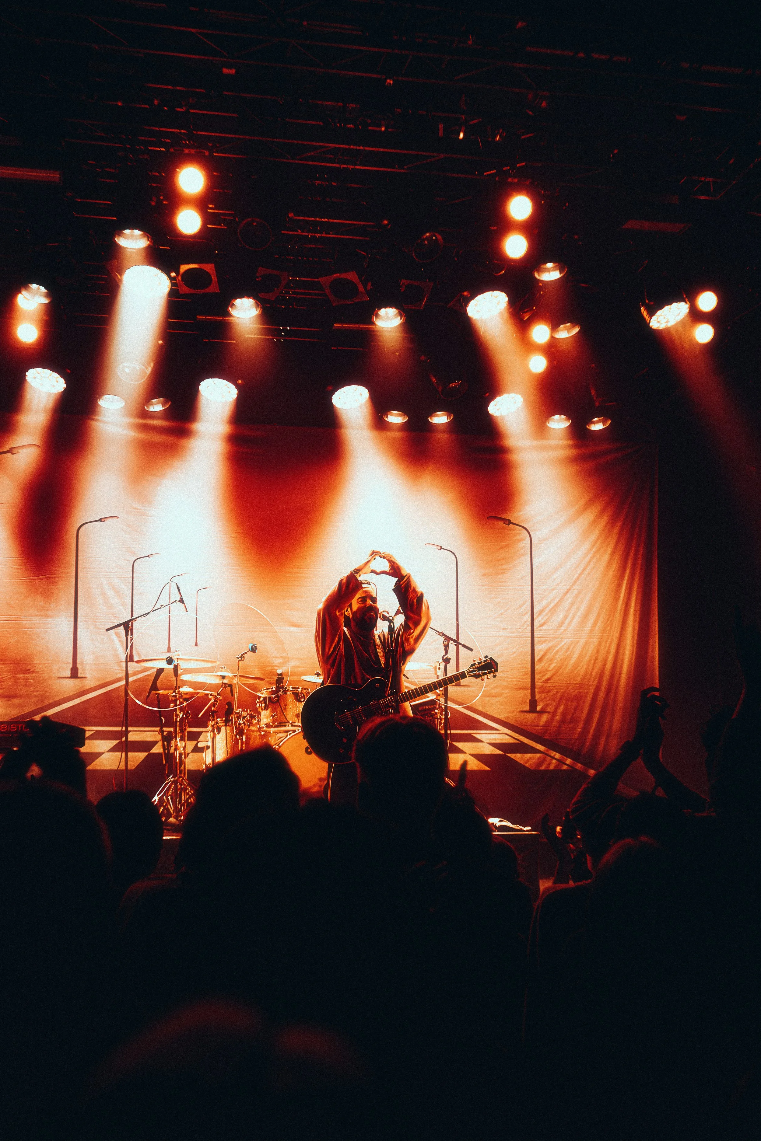 A musician standing on stage with a guitar, performing at a concert with stage lights and a backdrop depicting a road with streetlights.