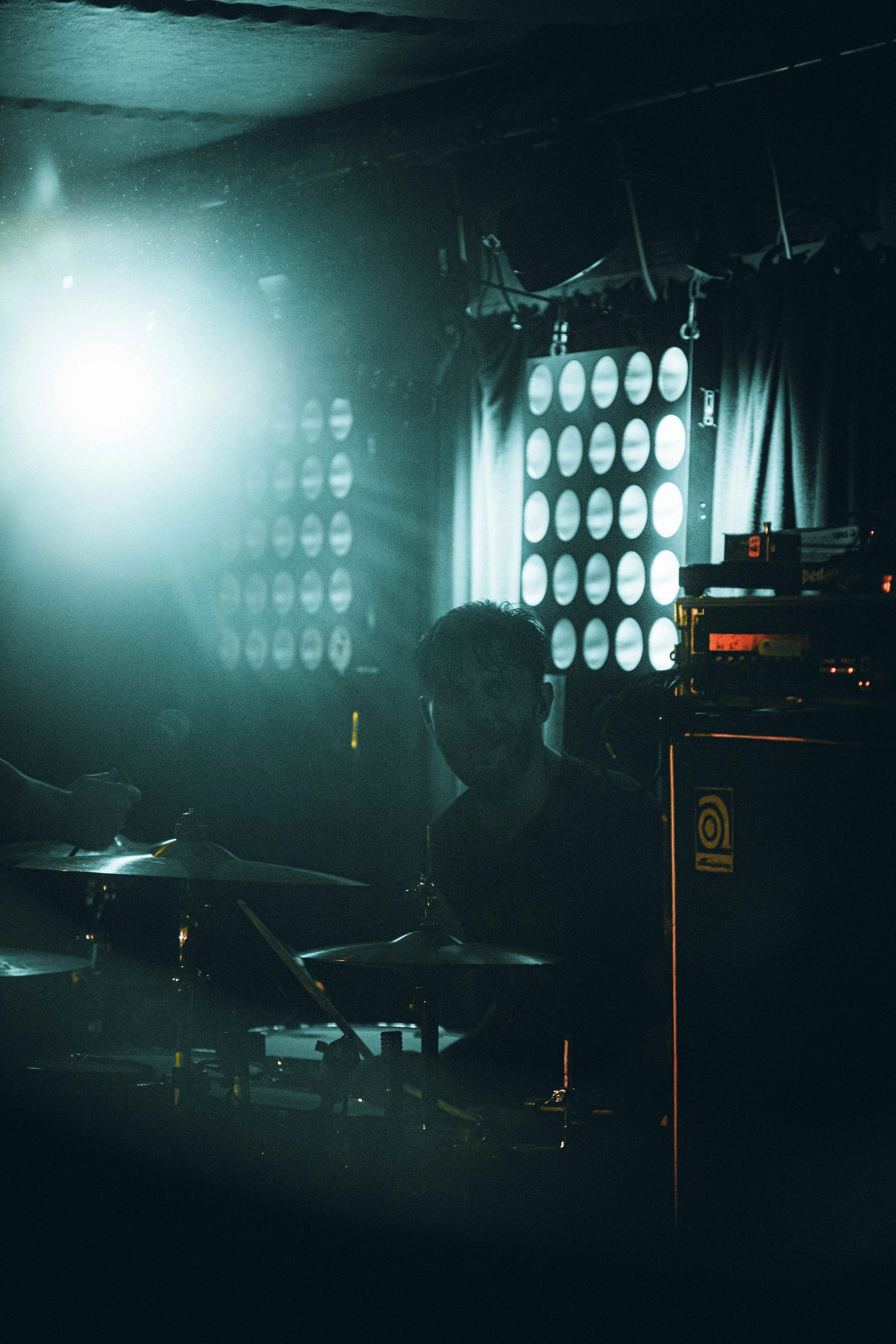 A person playing the drums on stage during a performance, with stage lighting and equipment visible in the background.