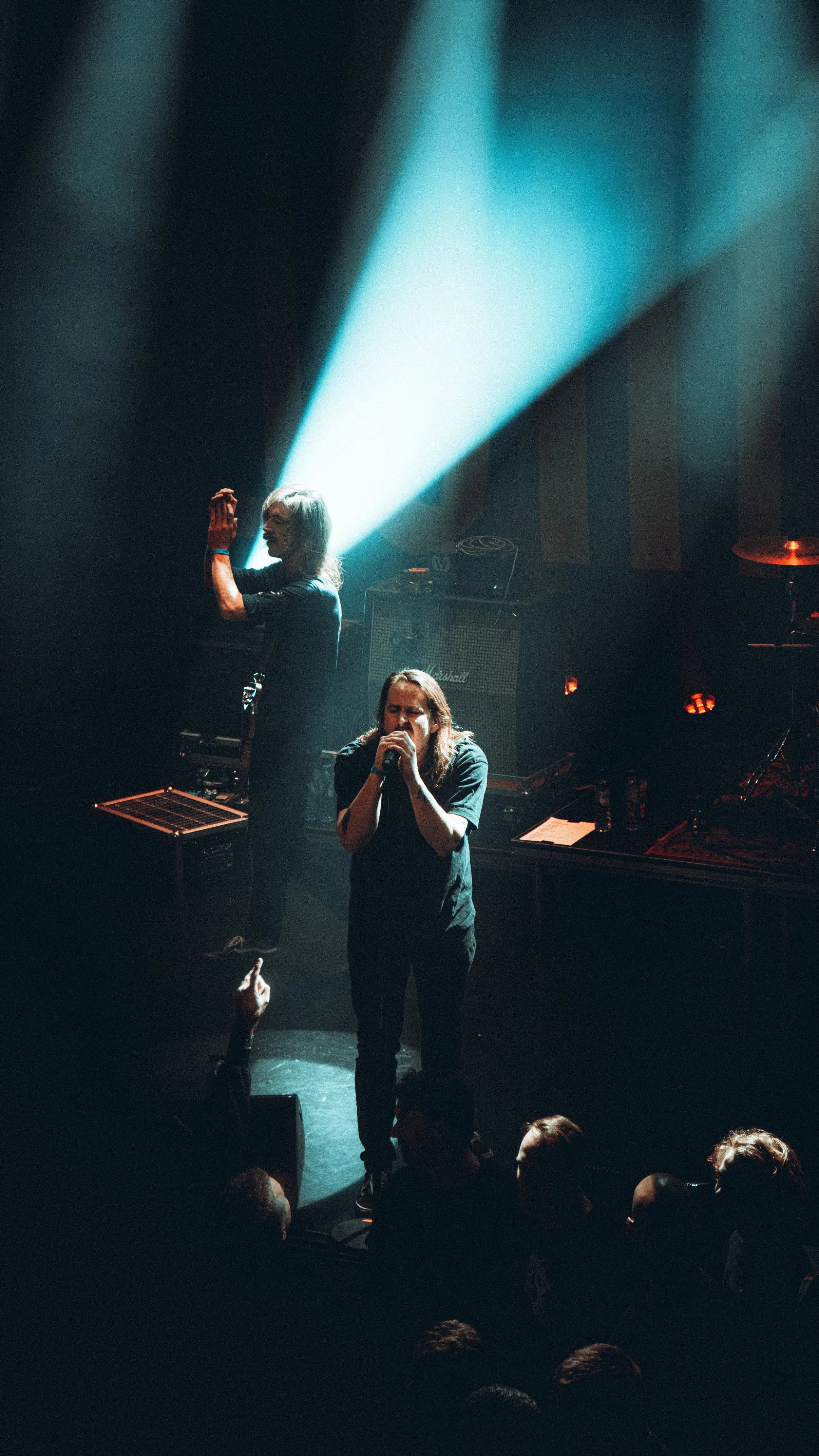 A live concert scene with a singer holding a microphone, performing on stage, with another person standing behind and a spotlight shining down. Audience members are visible in the foreground.