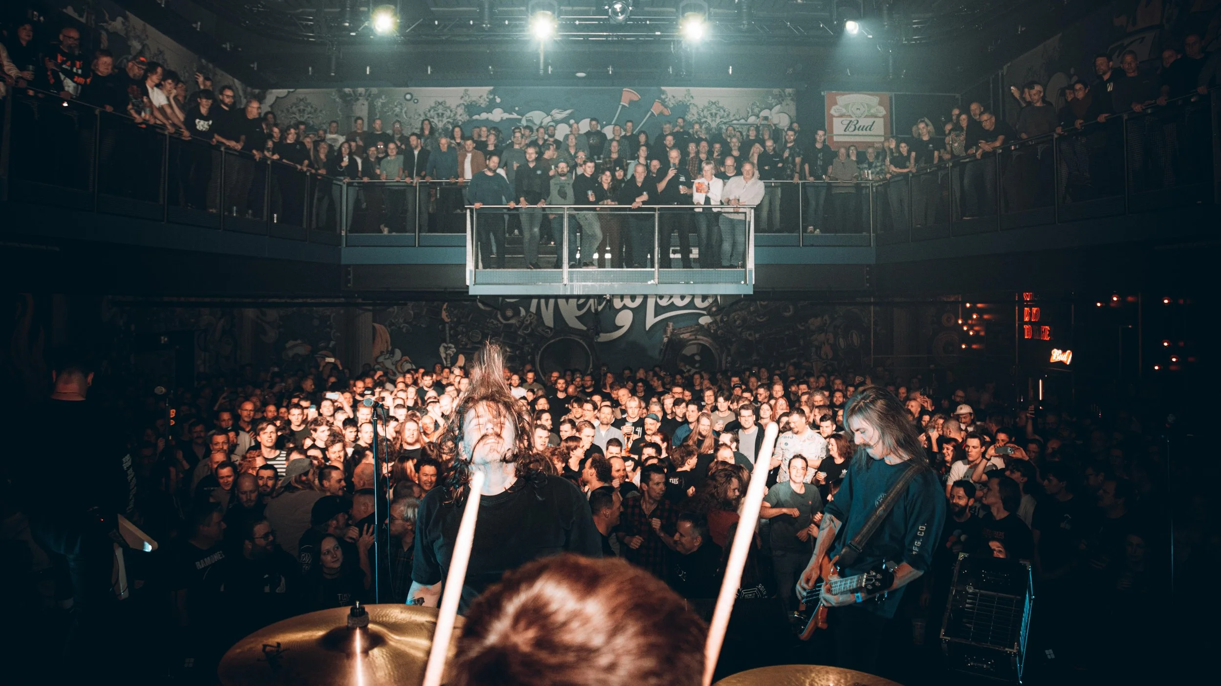 A live music concert with a large crowd, band members performing on stage, and a second level with audience members overlooking the performance.