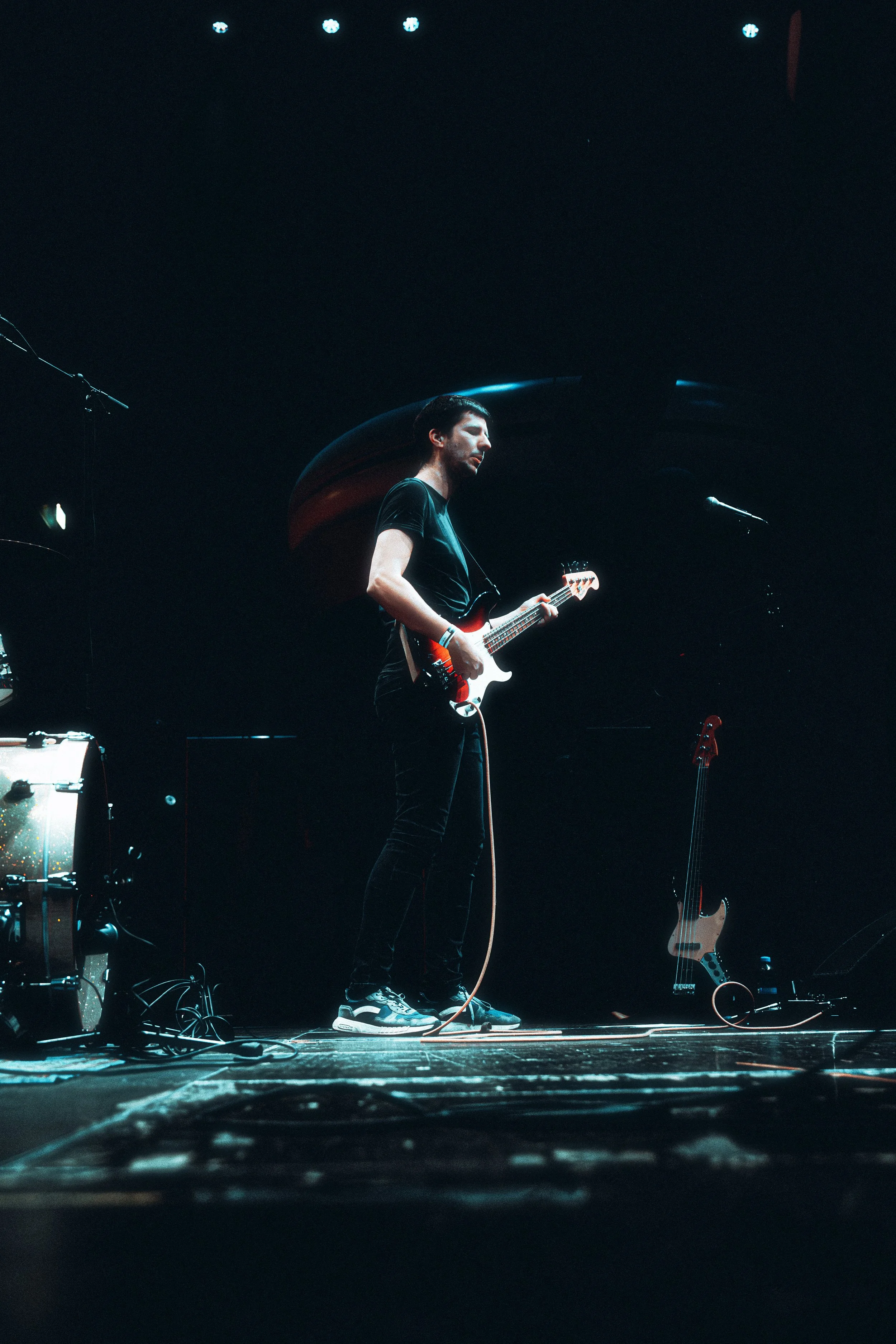 A male guitarist performing on a dark stage, playing an electric guitar with a black background and stage lights.