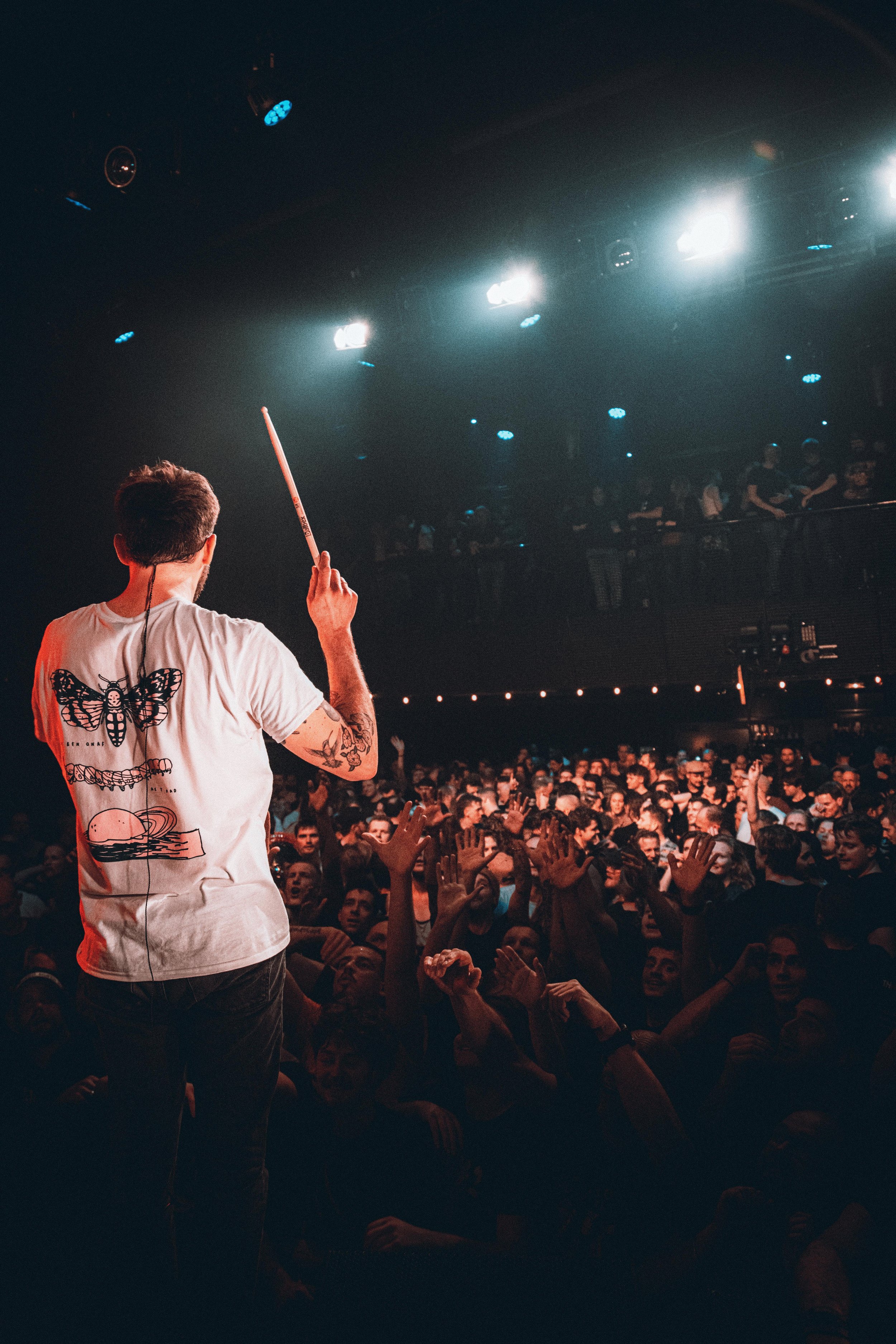 Front view of a musician standing on stage with his back to the camera, holding a drumstick, facing a large crowd at a concert venue with bright stage lights.