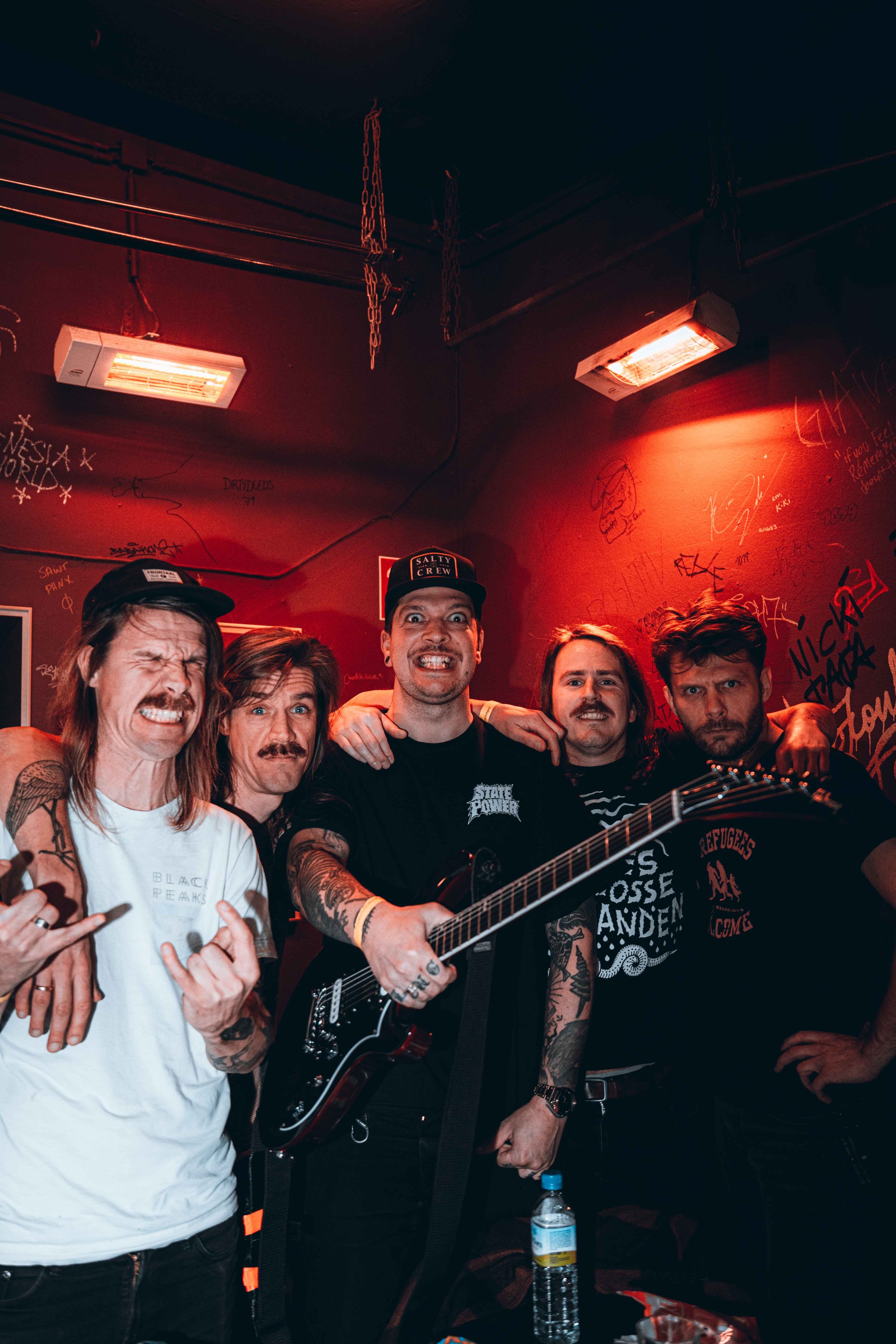 Group of five men posing together, one holding a guitar, in a red-lit room with graffiti on the walls.