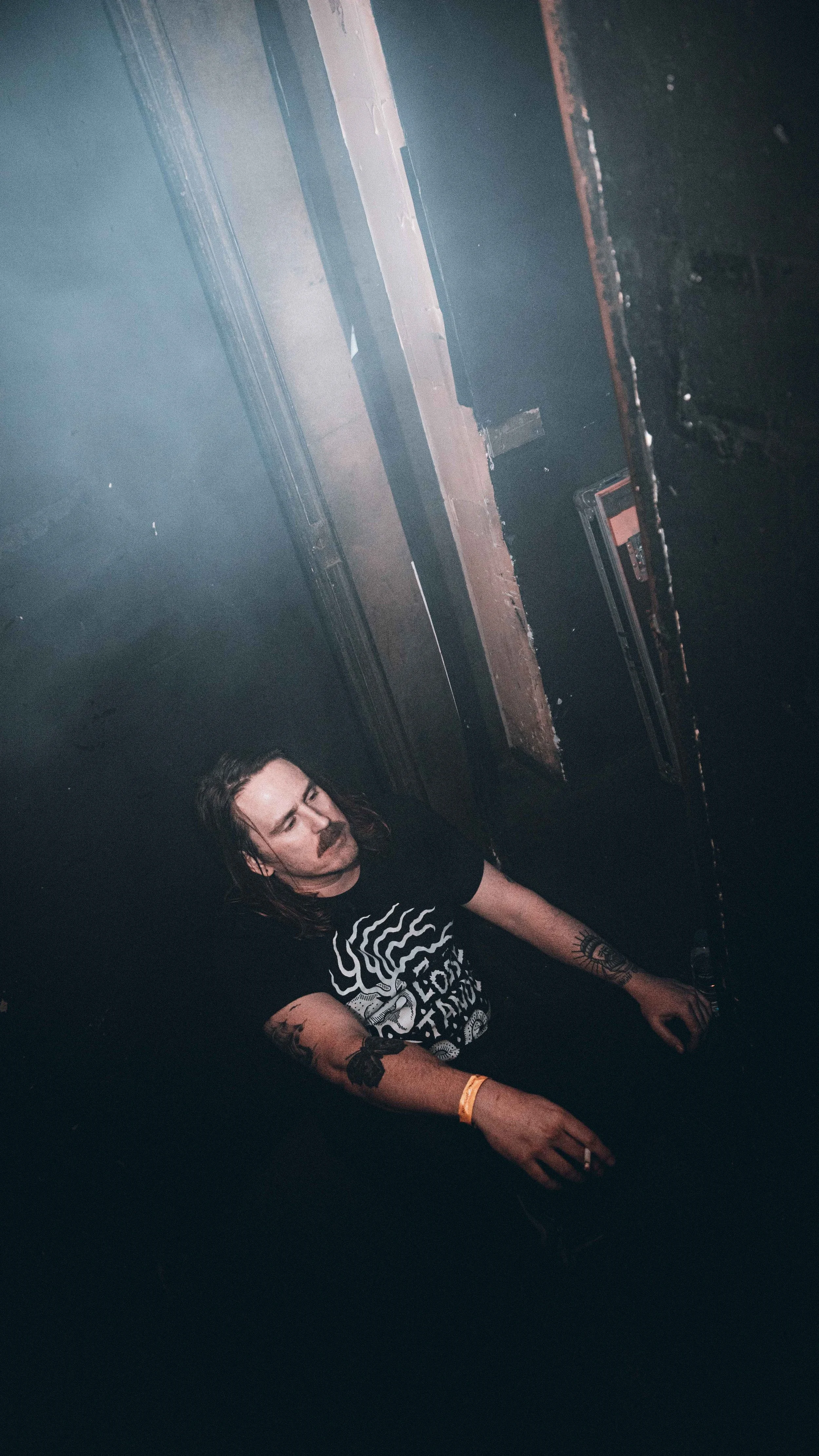 A man with long hair and tattoos on both arms is standing in front of a DJ mixer or sound equipment in a dark, smoky environment, possibly a club or music venue.