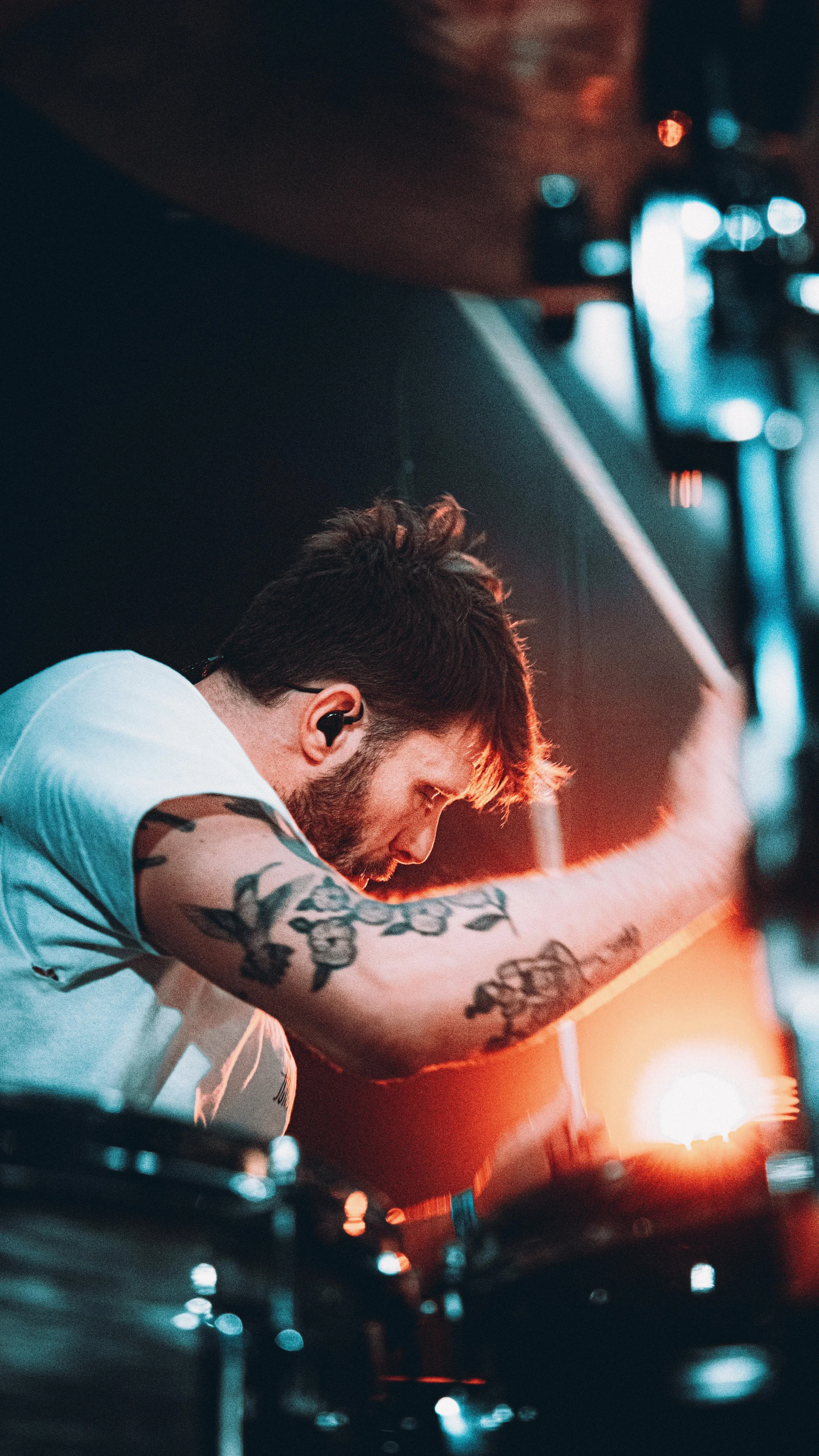 A male DJ with tattoos on his arm is focused on his equipment at a nightclub with colorful lighting.