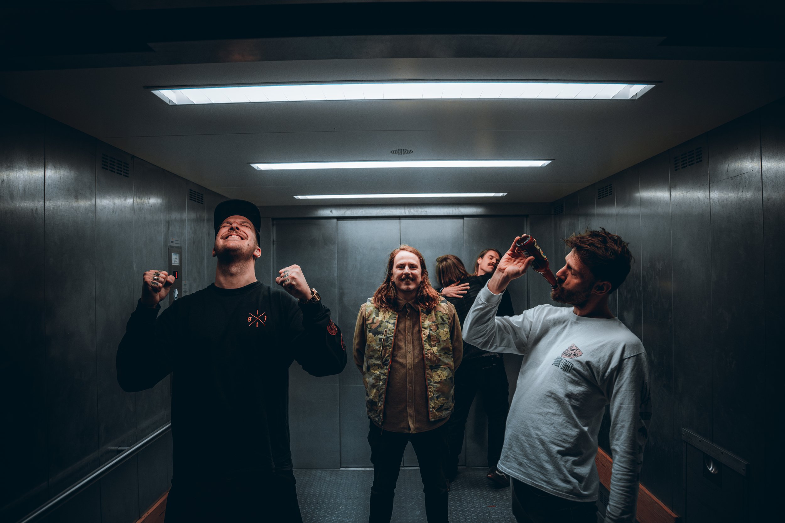 Group of five friends celebrating in elevator, some hugging, one drinking beer, others smiling with happiness.