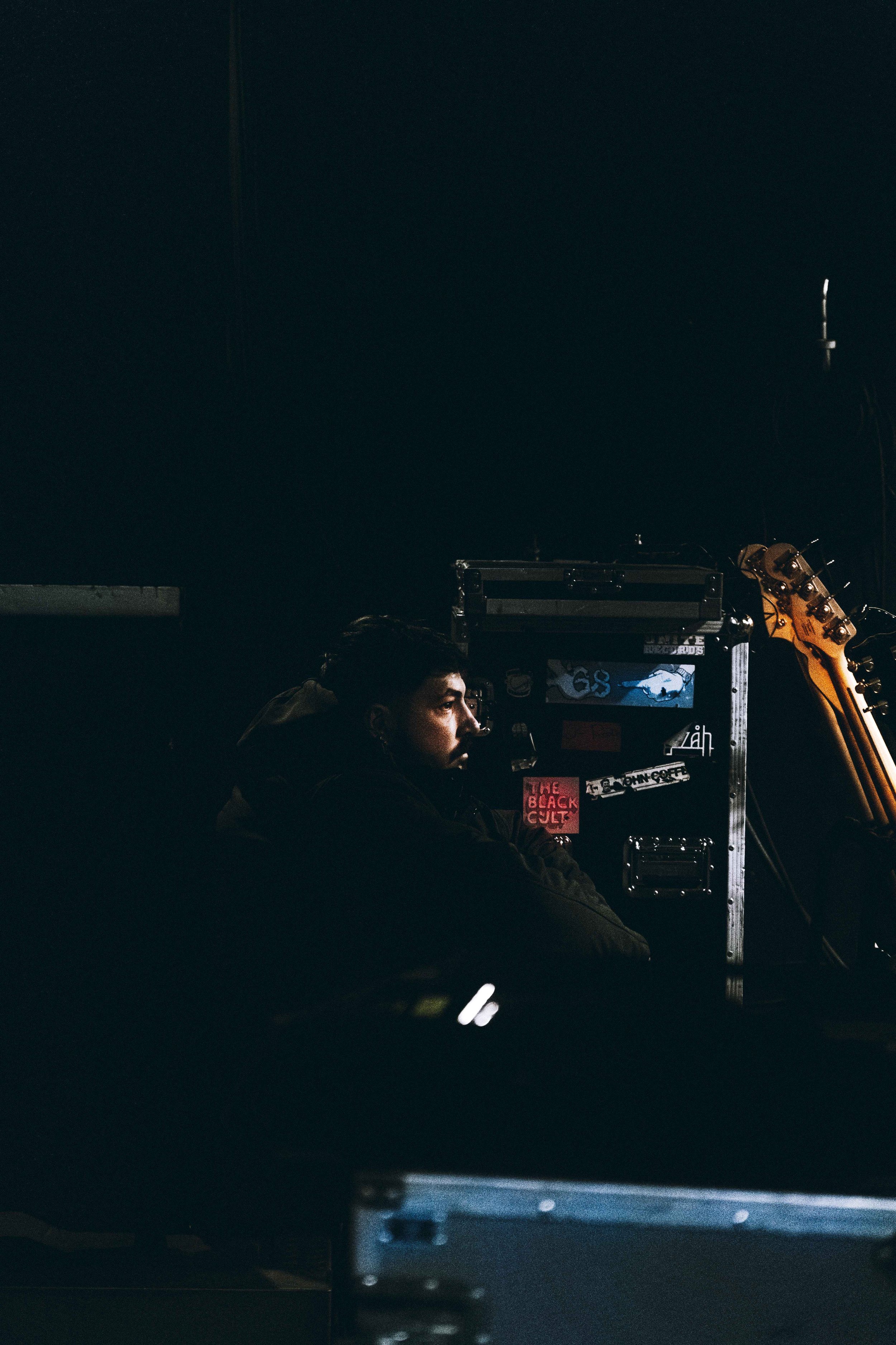 Man sitting in a dark room, partially illuminated, with sound equipment and a guitar nearby.