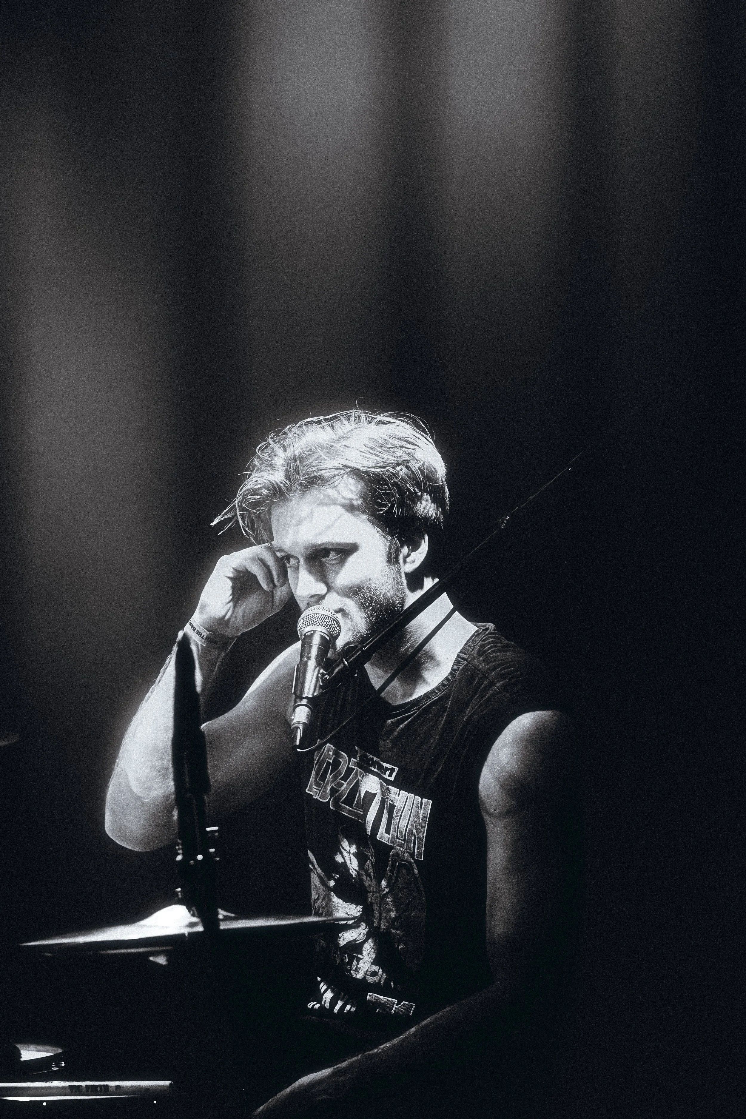 A black and white photo of a young man with dark, tousled hair, sitting at a piano with a microphone in front of him. He is wearing a sleeveless Led Zeppelin T-shirt and appears to be deep in thought during a performance.