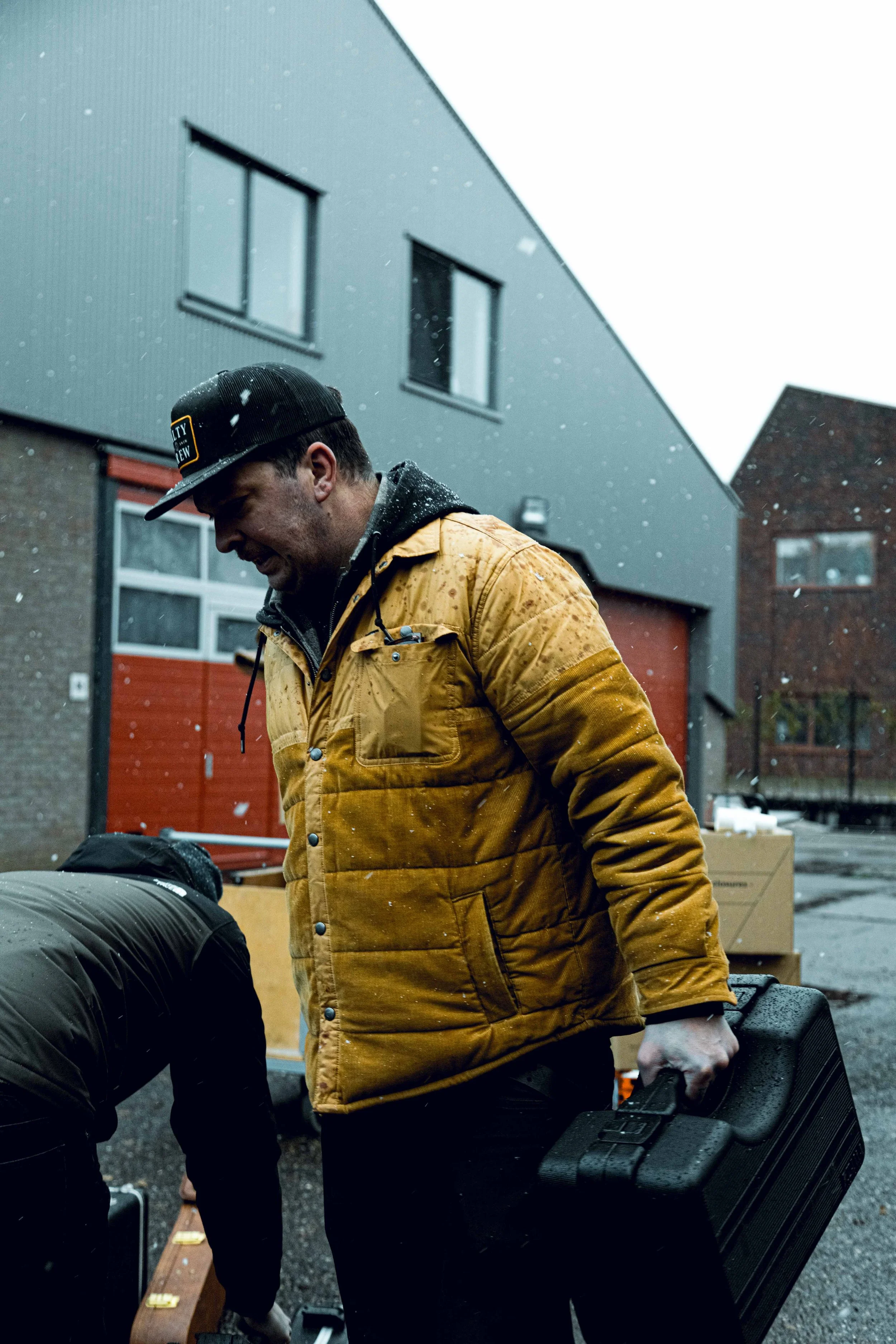 A man in a yellow jacket holding a black toolbox, standing outdoors on a rainy day near modern buildings.