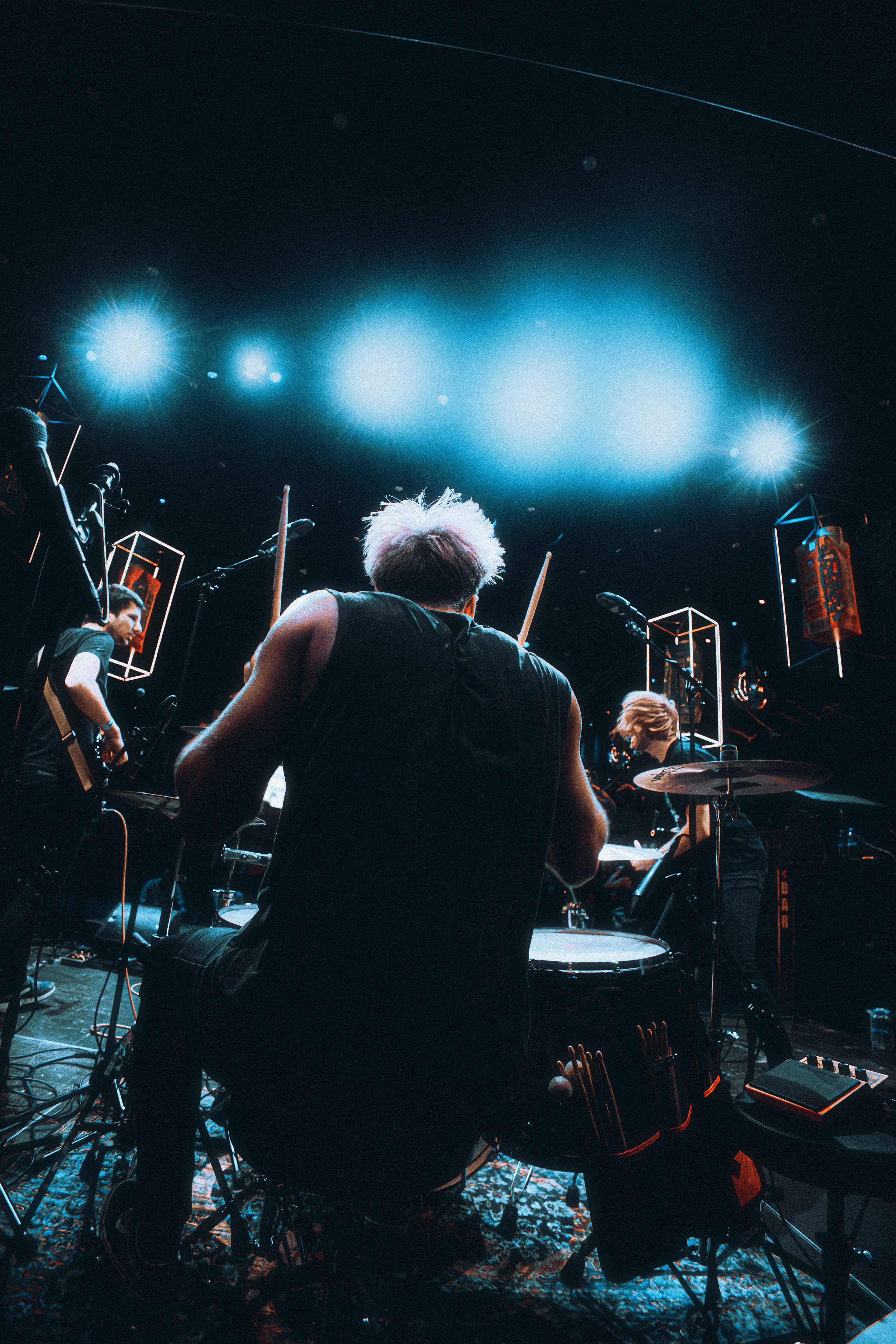 A band performing on stage with a drummer in the foreground, backlit with bright stage lights.