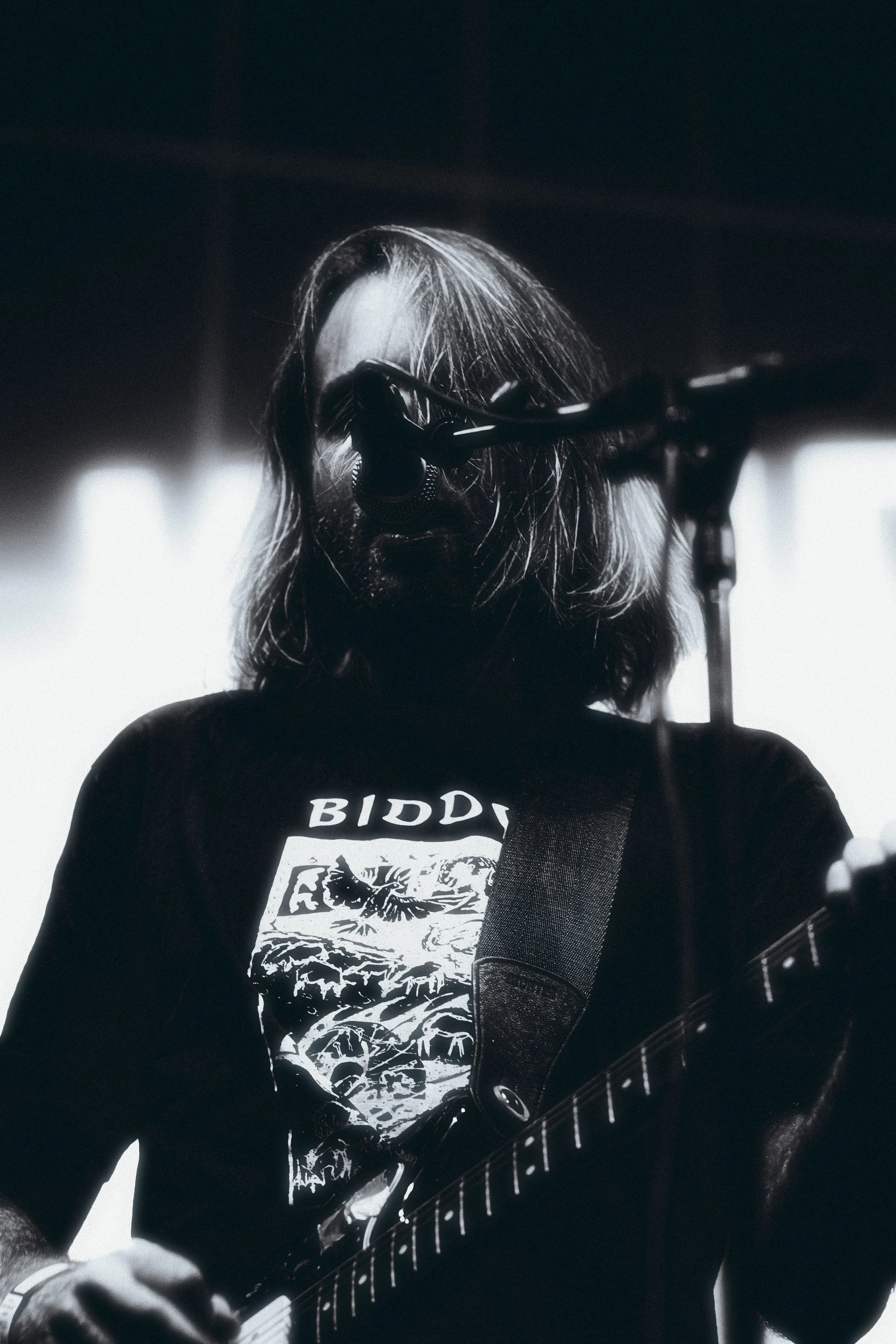 A male musician with long hair and a beard is playing an electric guitar near a microphone, with backlighting creating a silhouette effect.