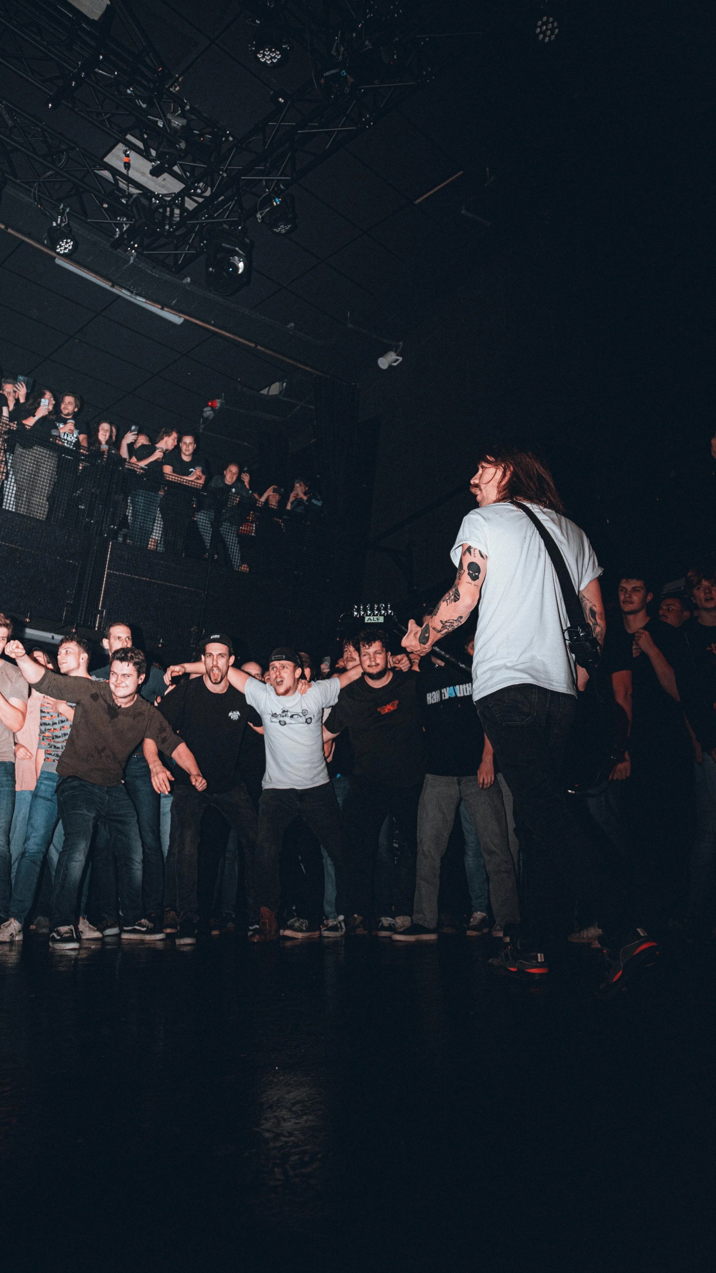 A performer with long hair and tattoos on their arms on stage, and a crowd of concertgoers standing close together, some with arms around each other, at a music concert in a dark indoor venue.