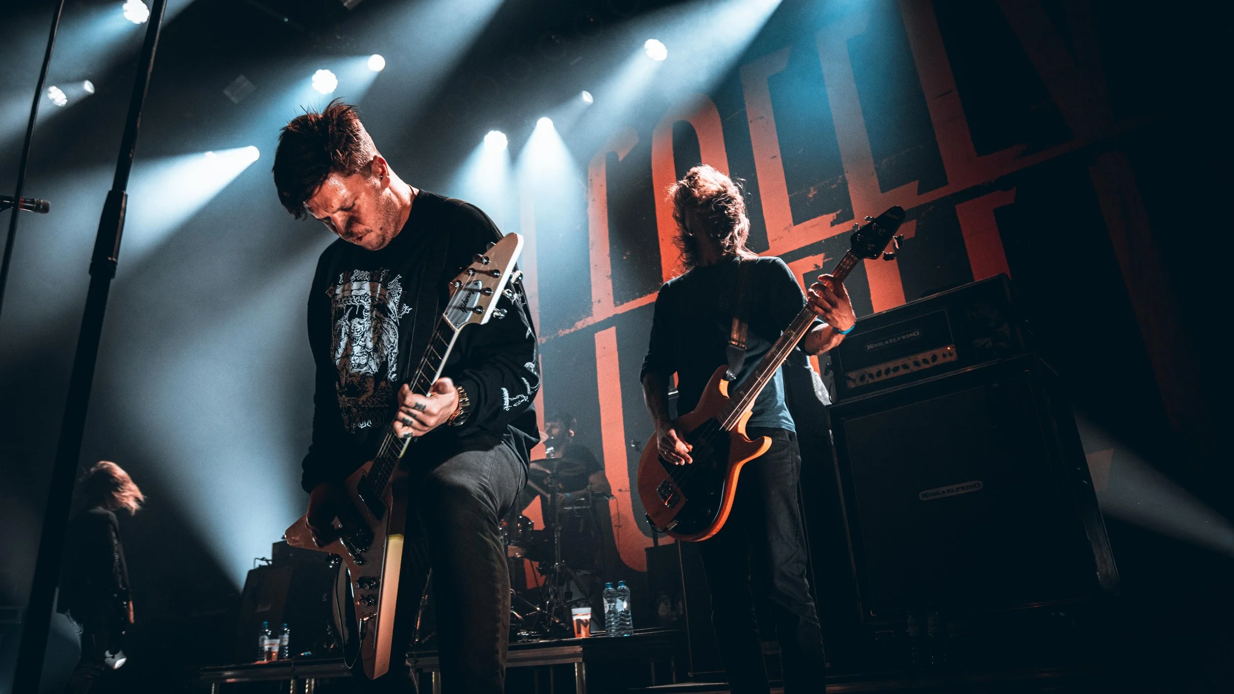 Two musicians performing on stage with guitars, with a large red and black backdrop and stage lights overhead.