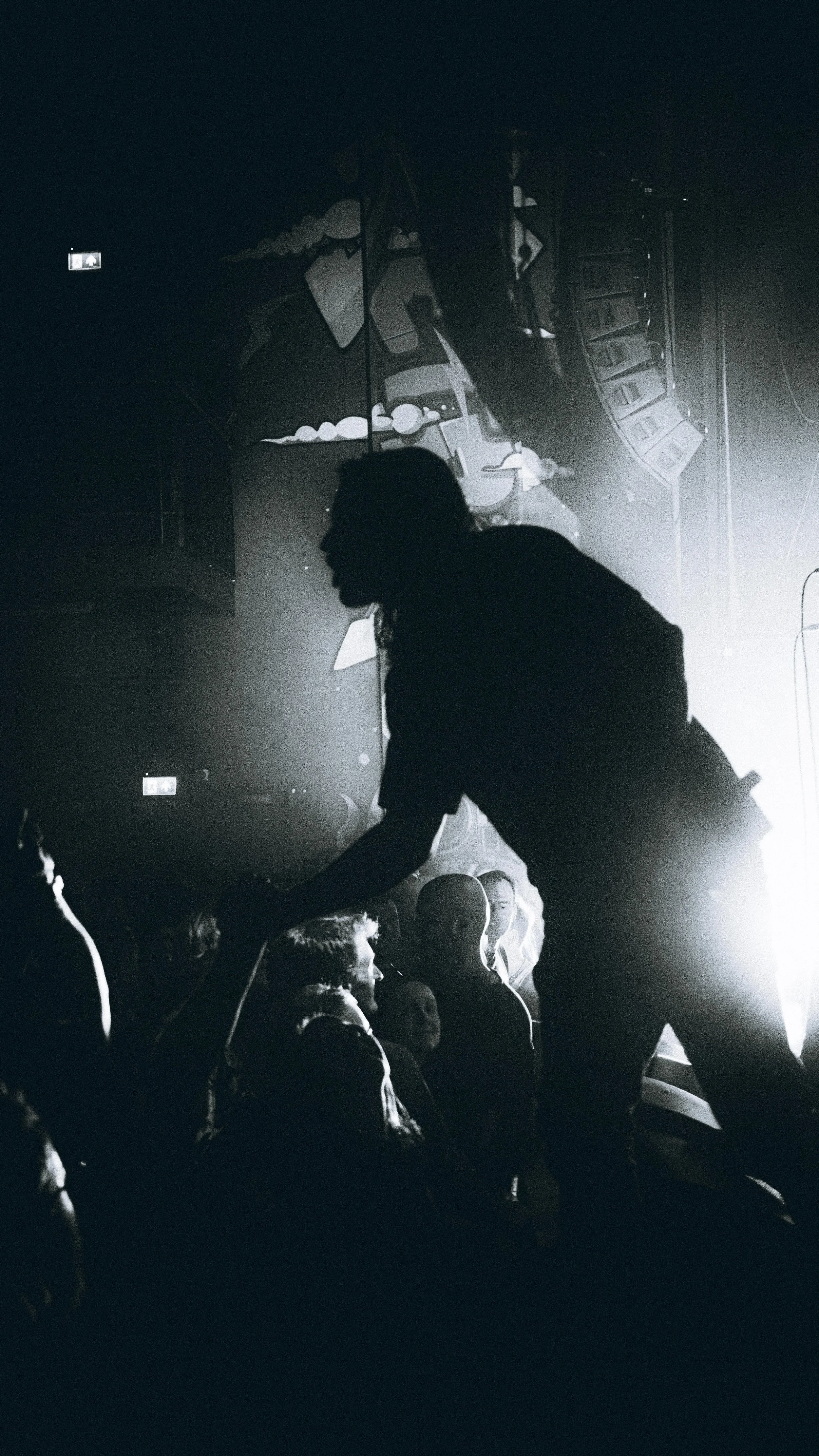 A silhouette of a person in black clothing reaching out to a group of people at a concert or event, with stage lights and speakers in the background.