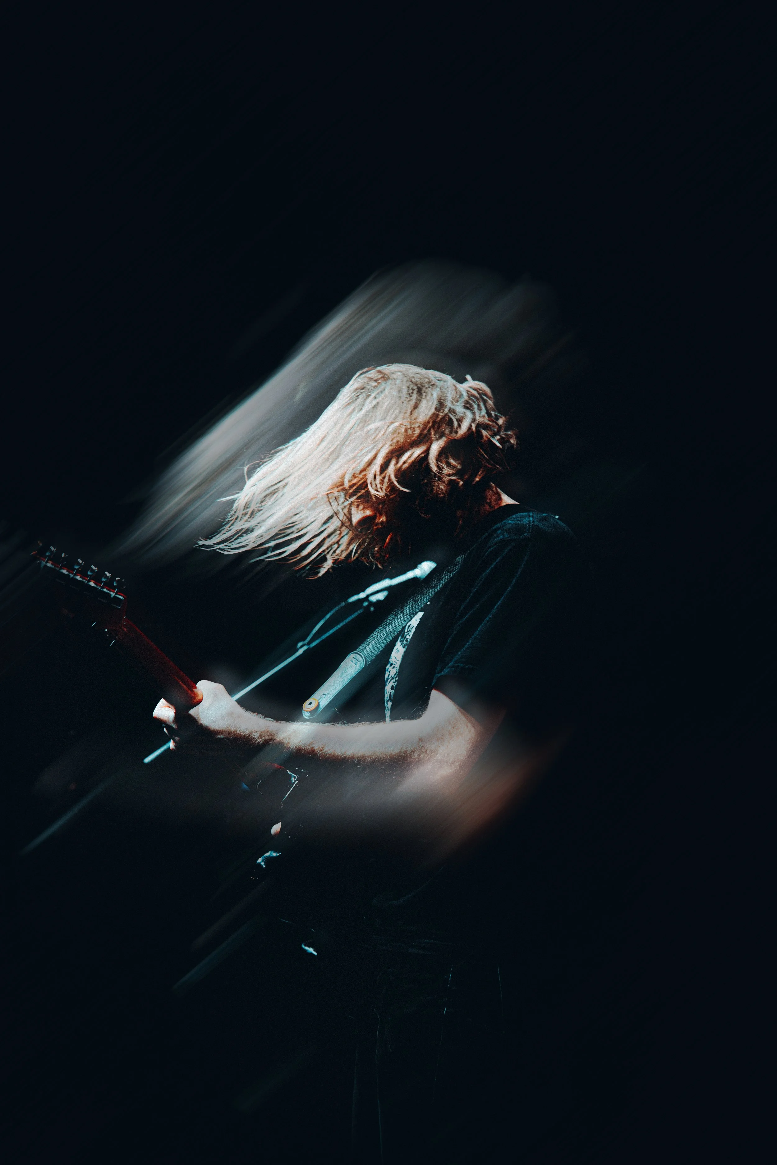 A person with long, blonde hair plays an electric guitar in a dark setting, with the photo captured using motion blur to create a dynamic effect.