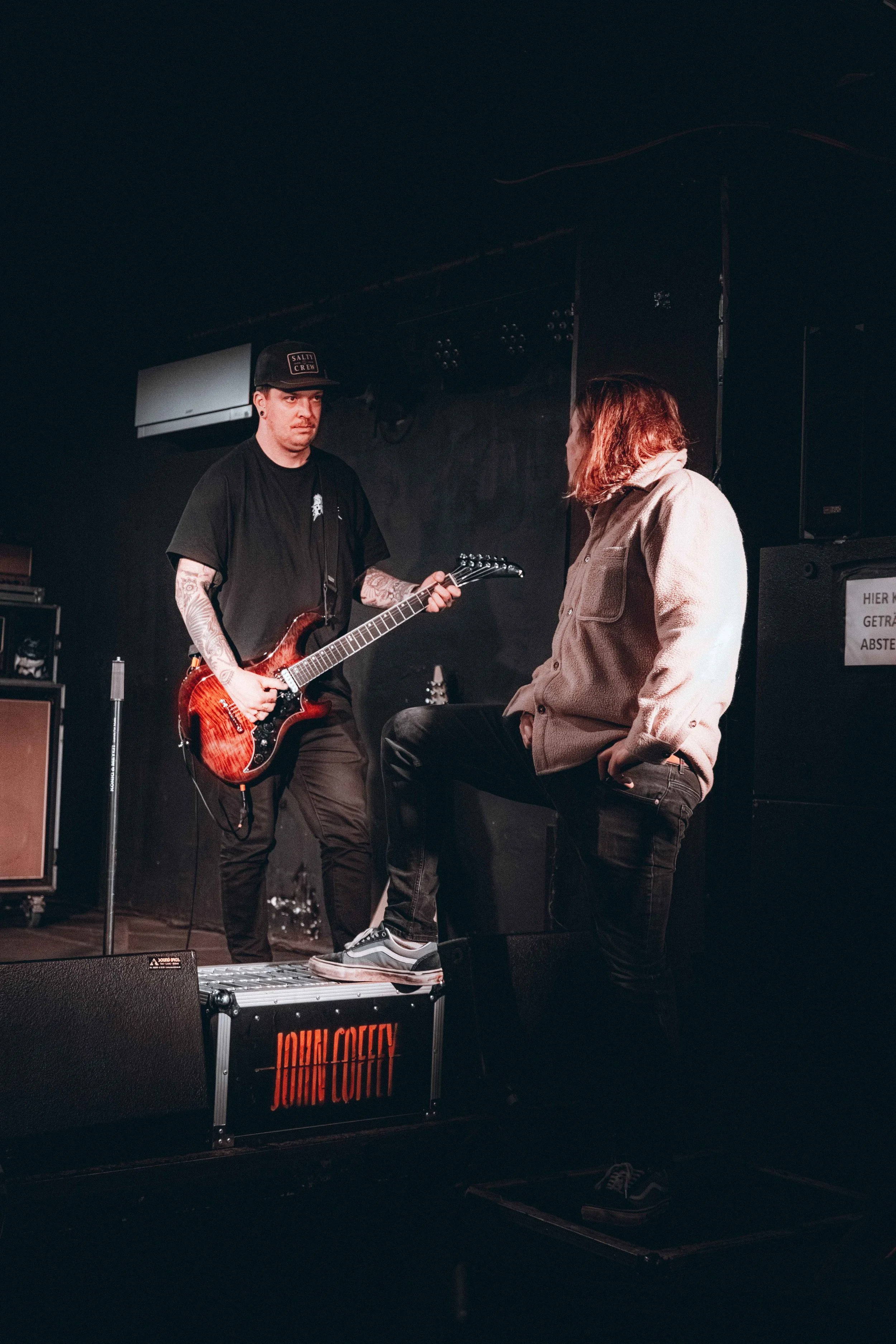 Two musicians on stage, one playing an electric guitar and the other standing with a foot on a monitor, engaged in a conversation or performance.