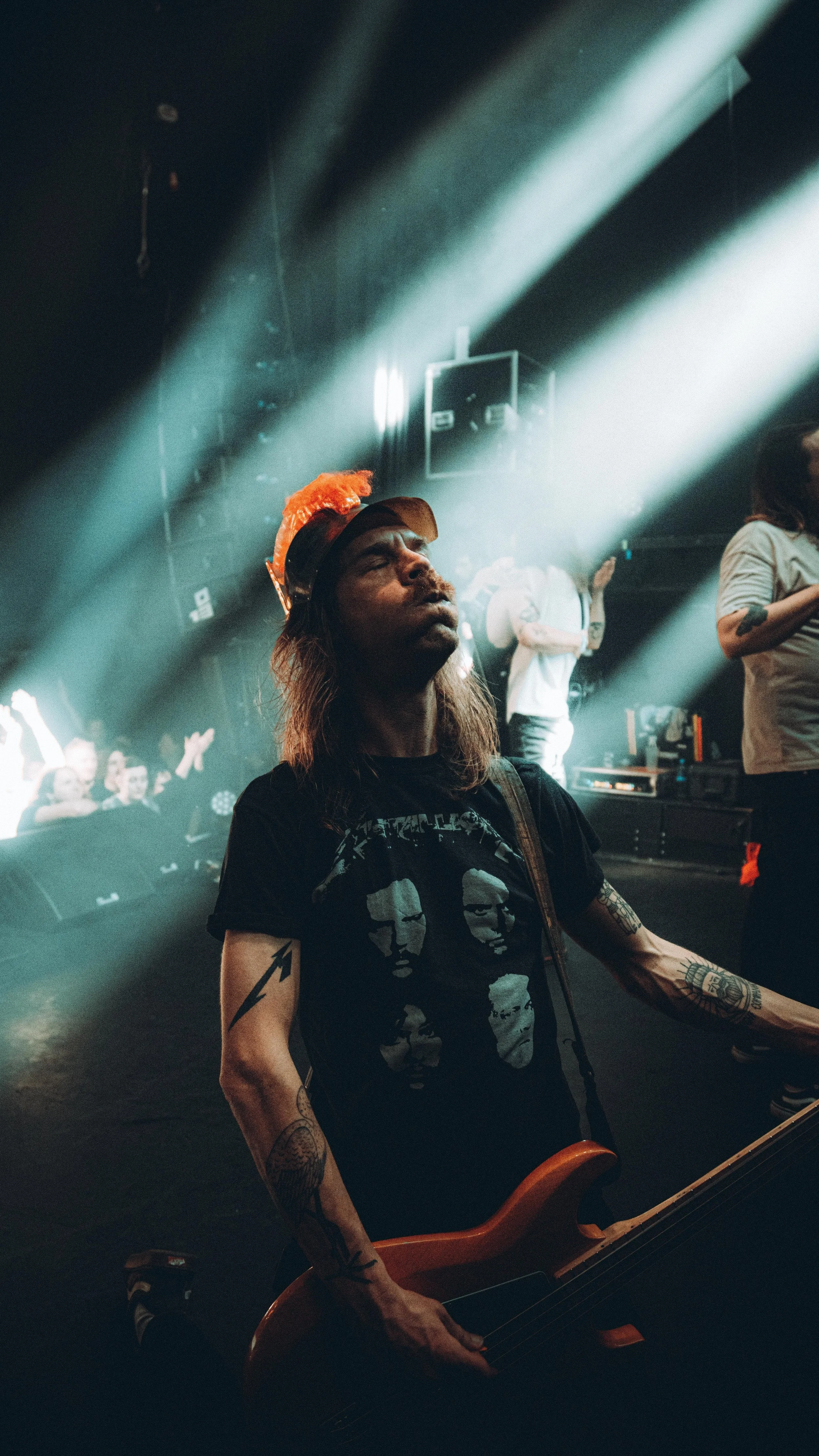A musician with tattoos on their arms playing a bass guitar under dramatic stage lighting at a concert.