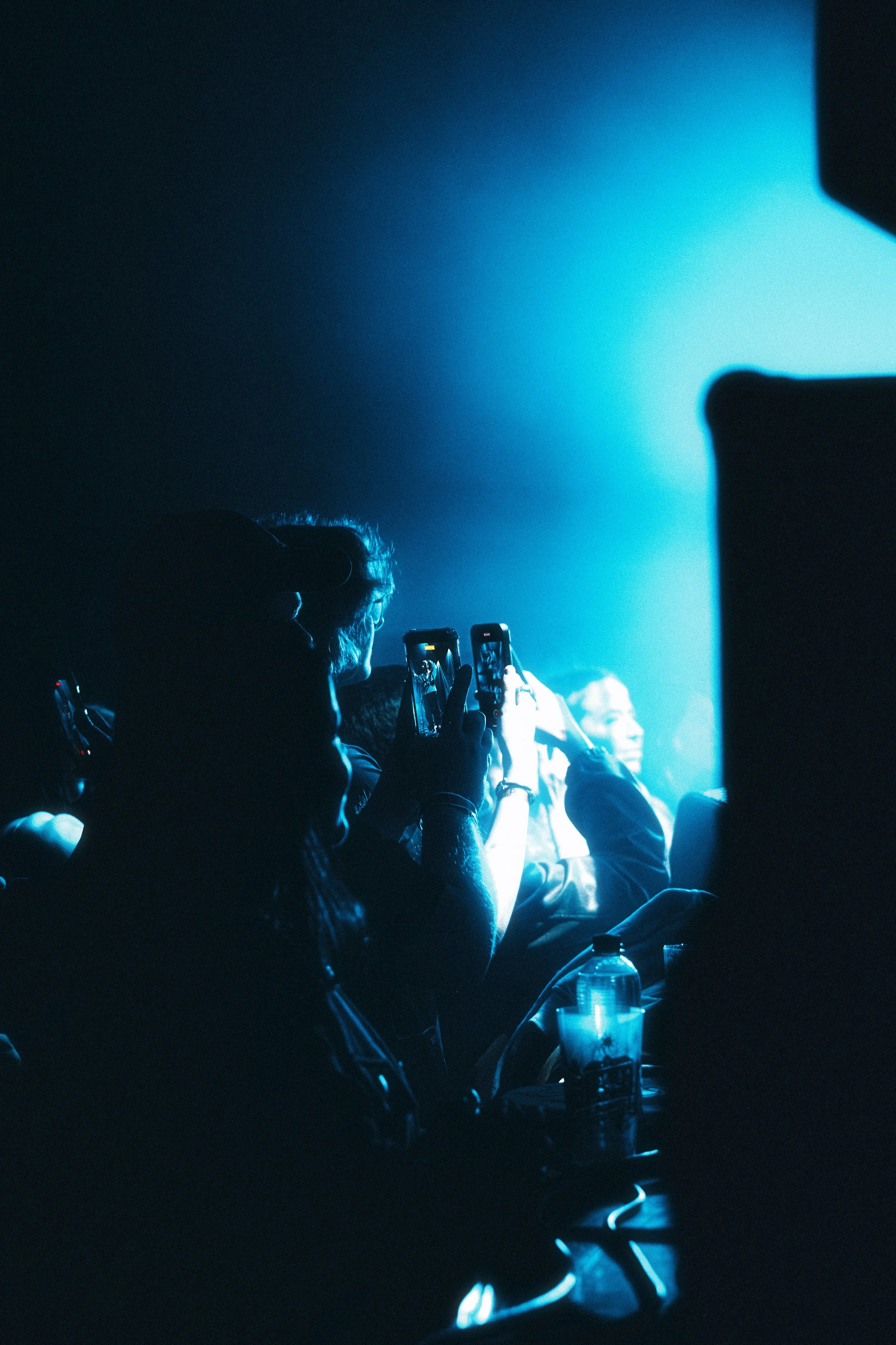 Audience members taking photos with their smartphones at a concert or event, illuminated by blue stage lighting.