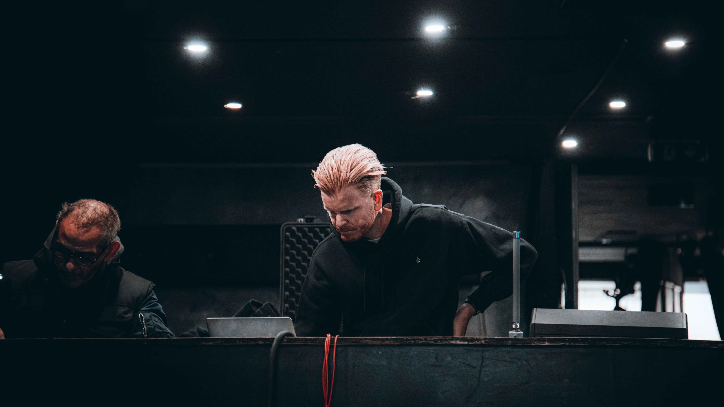 Man with pink hair leaning over a table in a dimly lit room, with another person sitting nearby