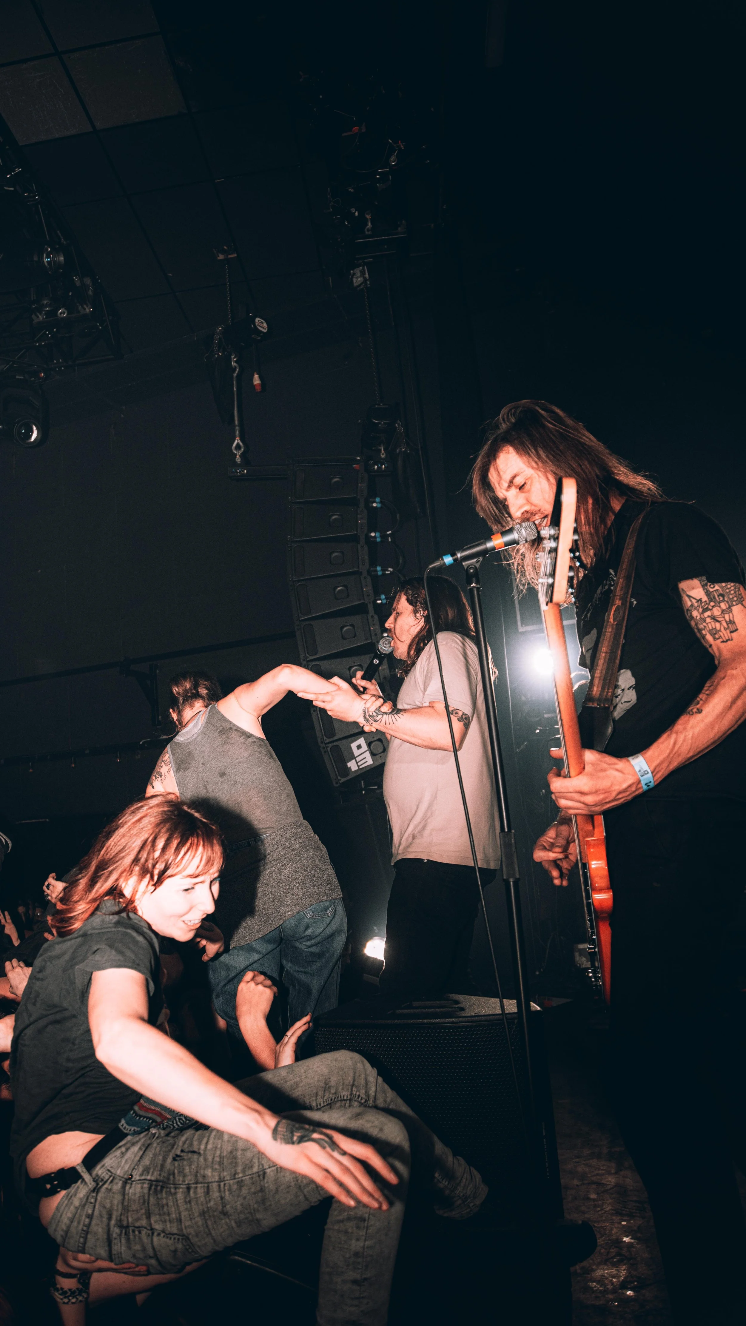 A rock band performs on stage with two singers, one of whom is crowd surfing, and a guitarist playing in the foreground at a concert.