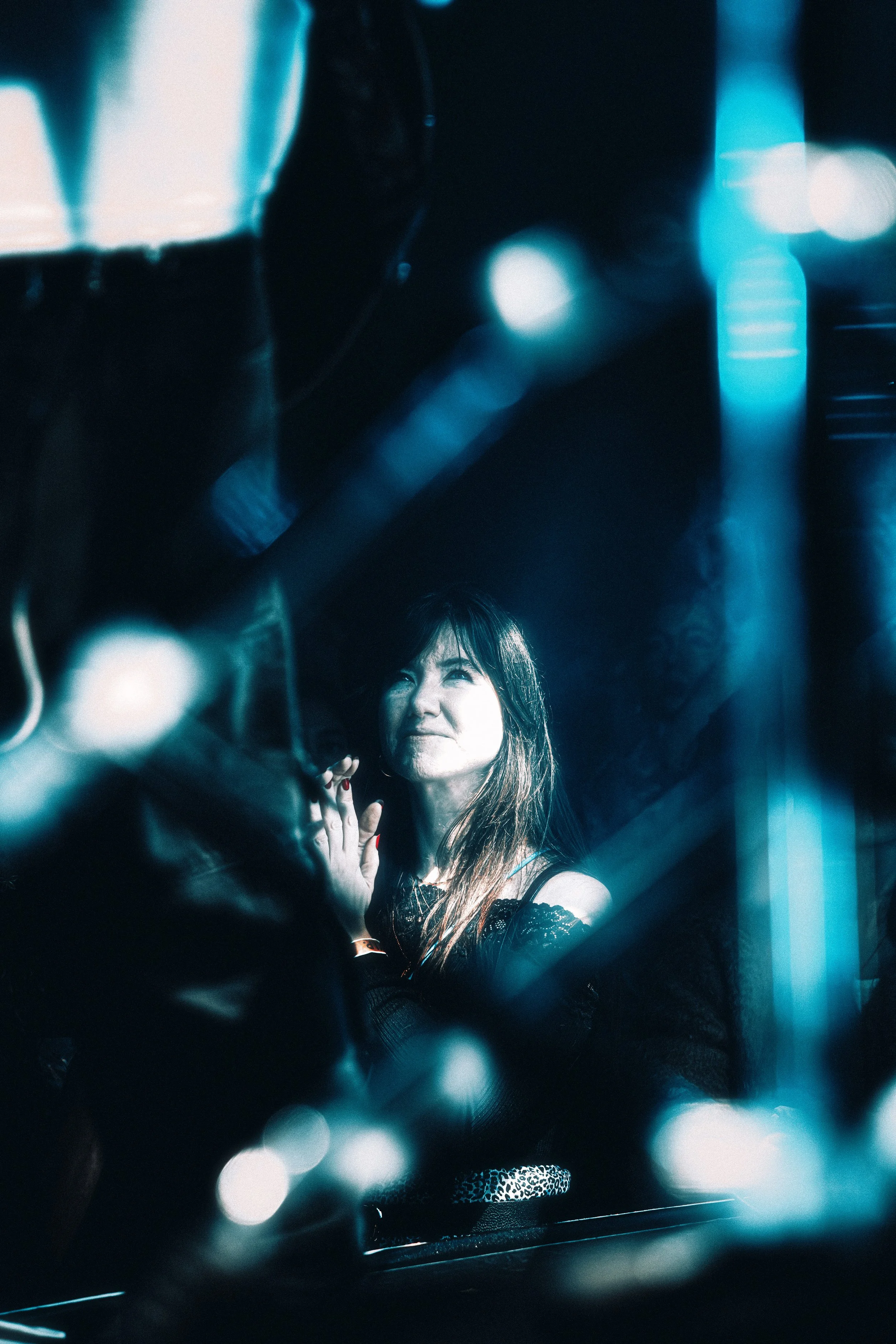A woman with long hair looking through a glass with a contemplative expression, illuminated by blue light, with blurred reflections and lights around her.
