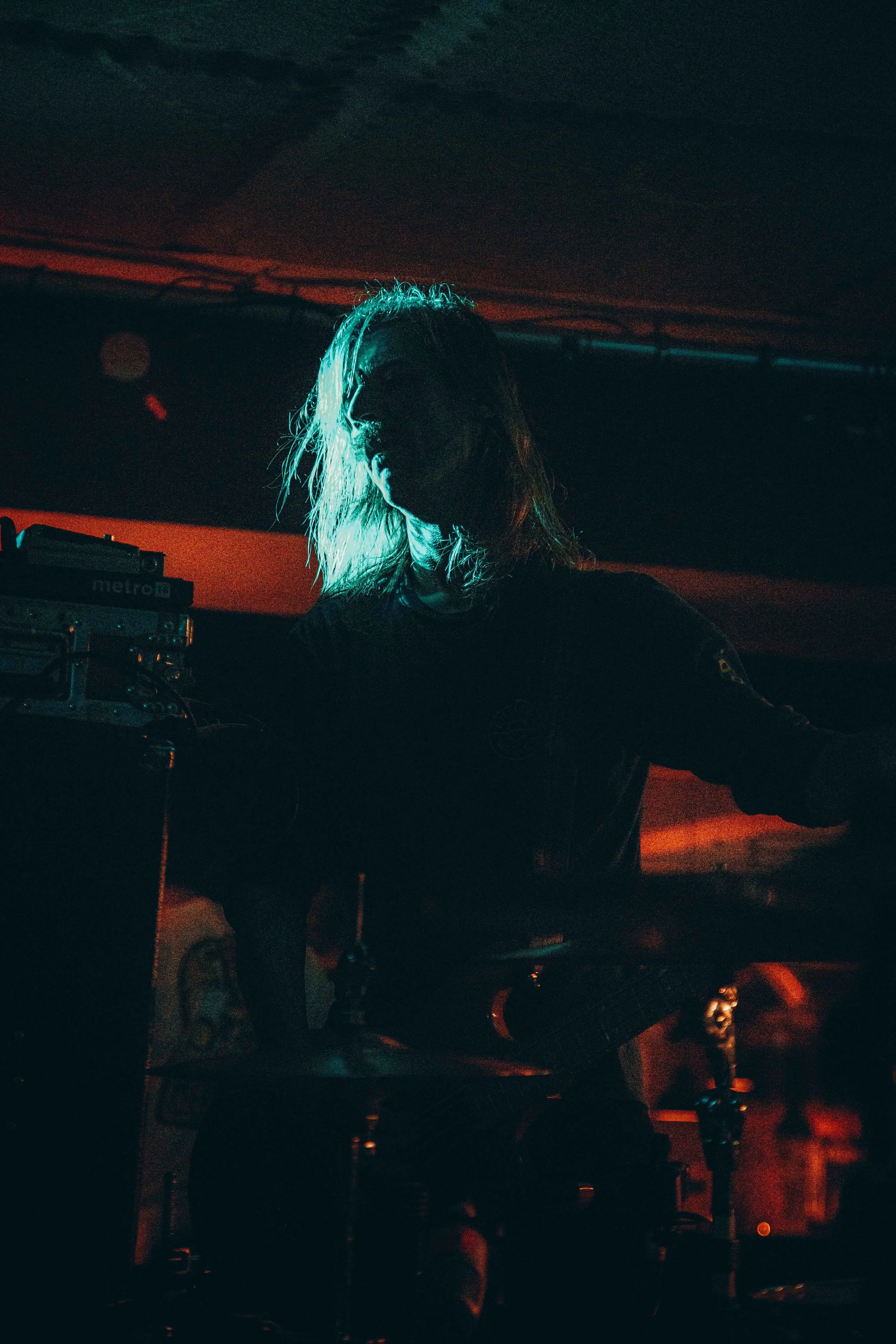 A person with long hair illuminated by blue and red stage lights, standing behind a drum set in a dark environment, possibly a concert or performance venue.
