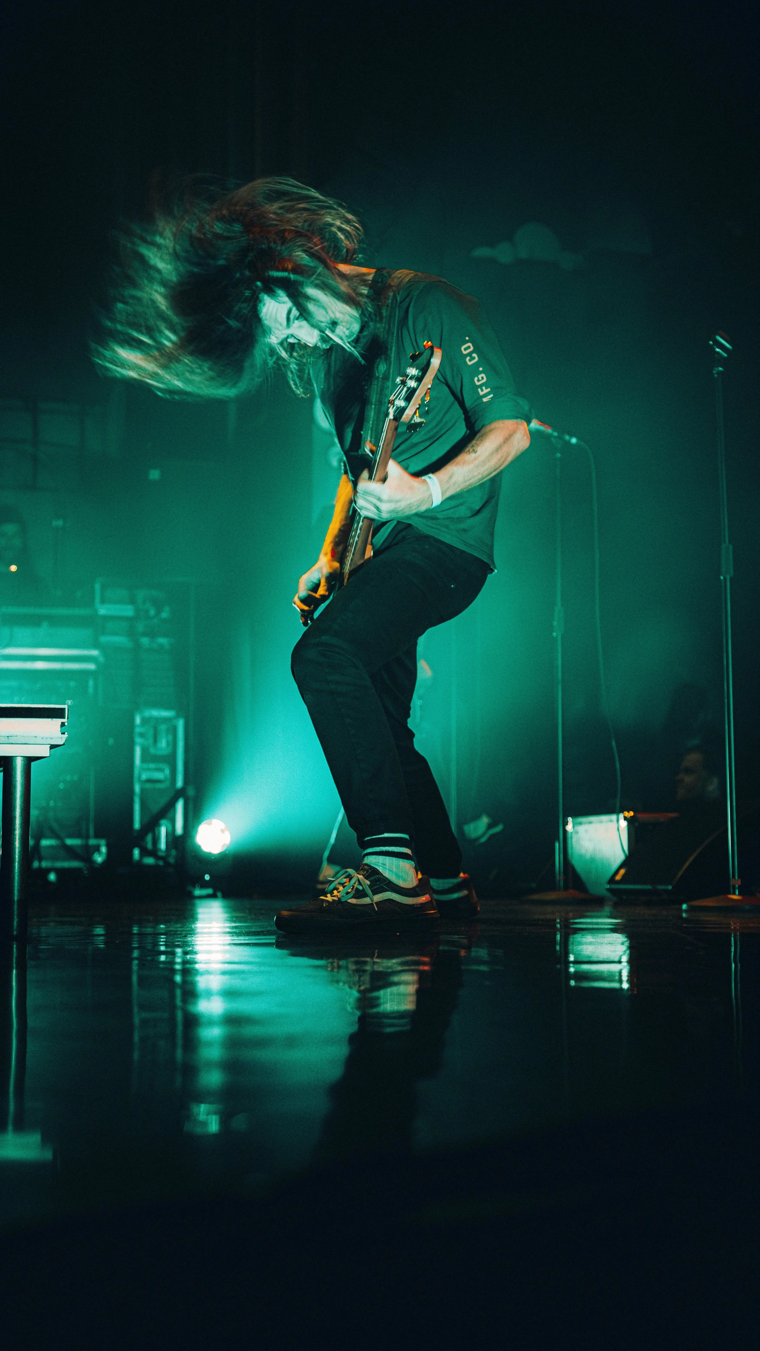Guitarist playing electric guitar on stage with long hair swinging, green/blue stage lighting.