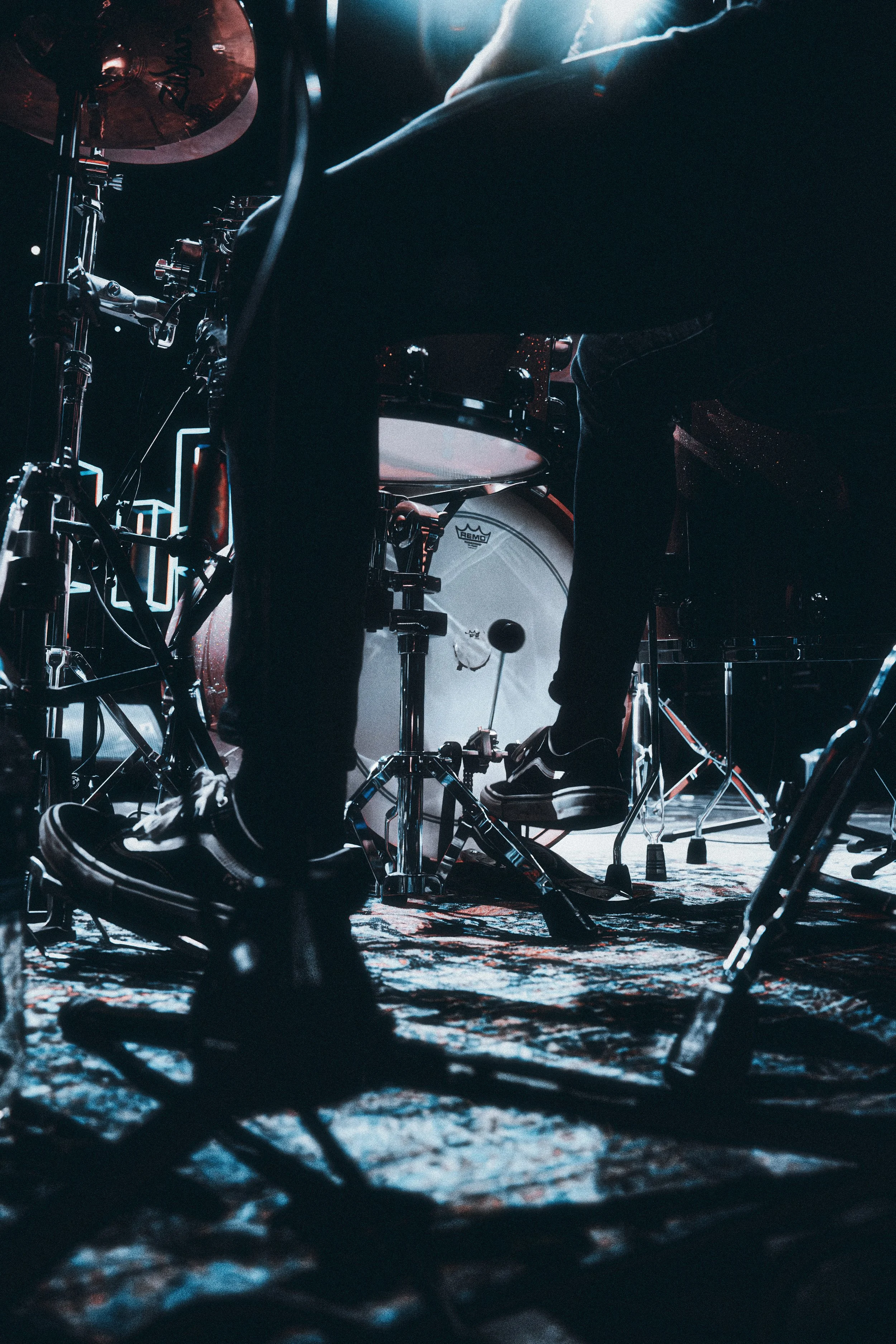 Close-up of a drummer playing a drum kit on stage, showing his legs and the drums from a low angle in a dimly lit setting with stage lights.