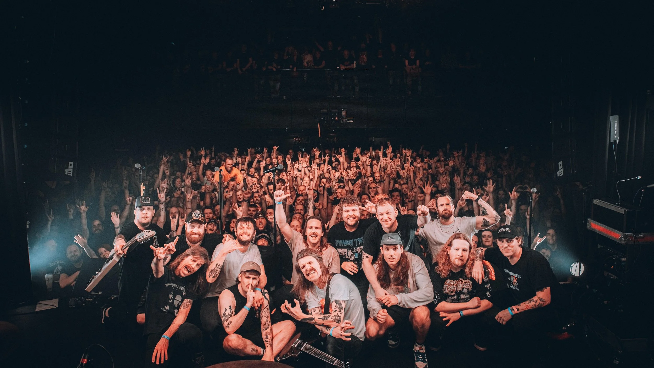 A large crowd of concert-goers raising their arms and cheering in a dark concert venue, with a group of band members posing and smiling in the foreground.
