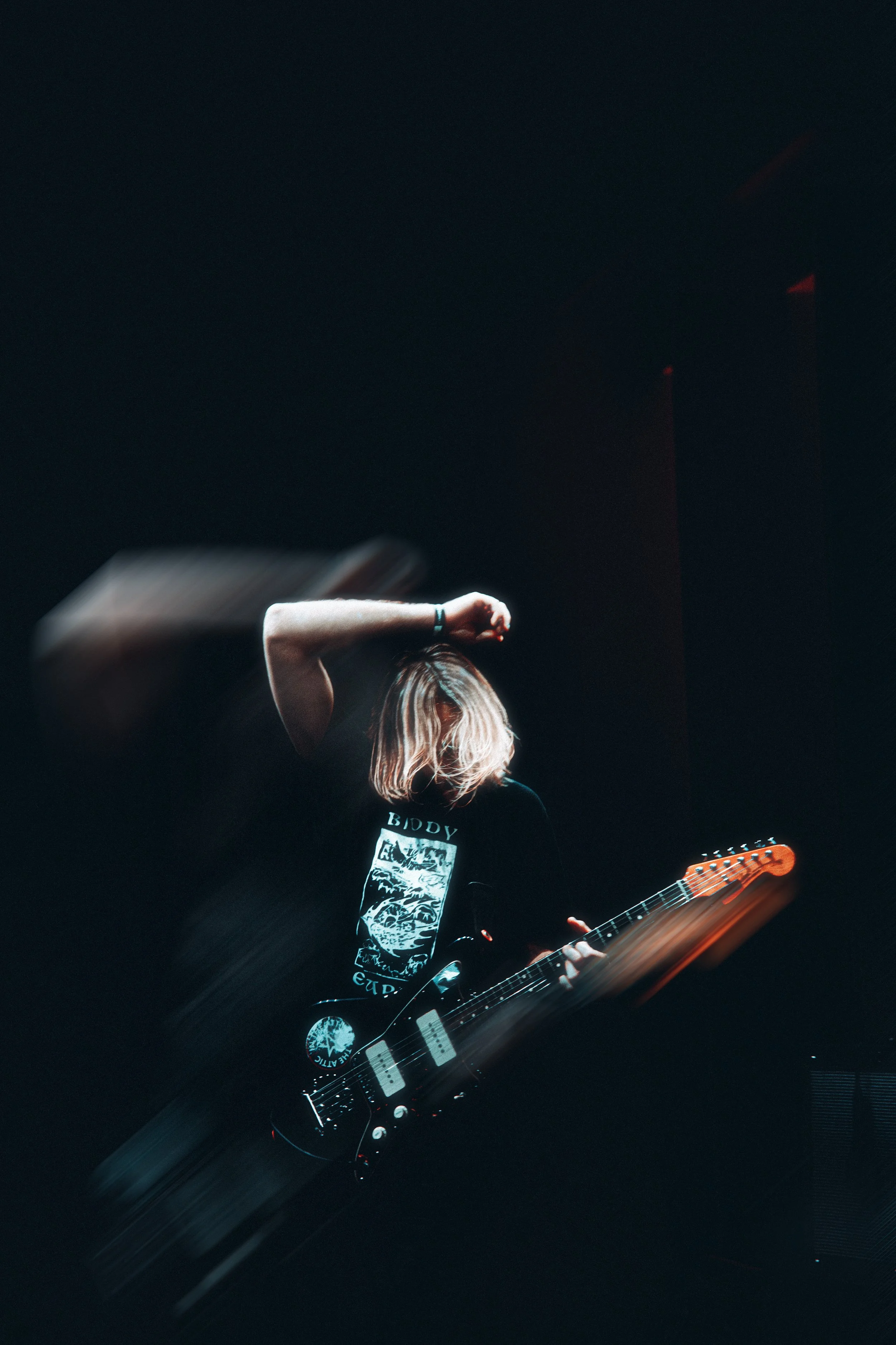 A person with blonde hair playing an electric guitar on stage with dark background and motion blur.