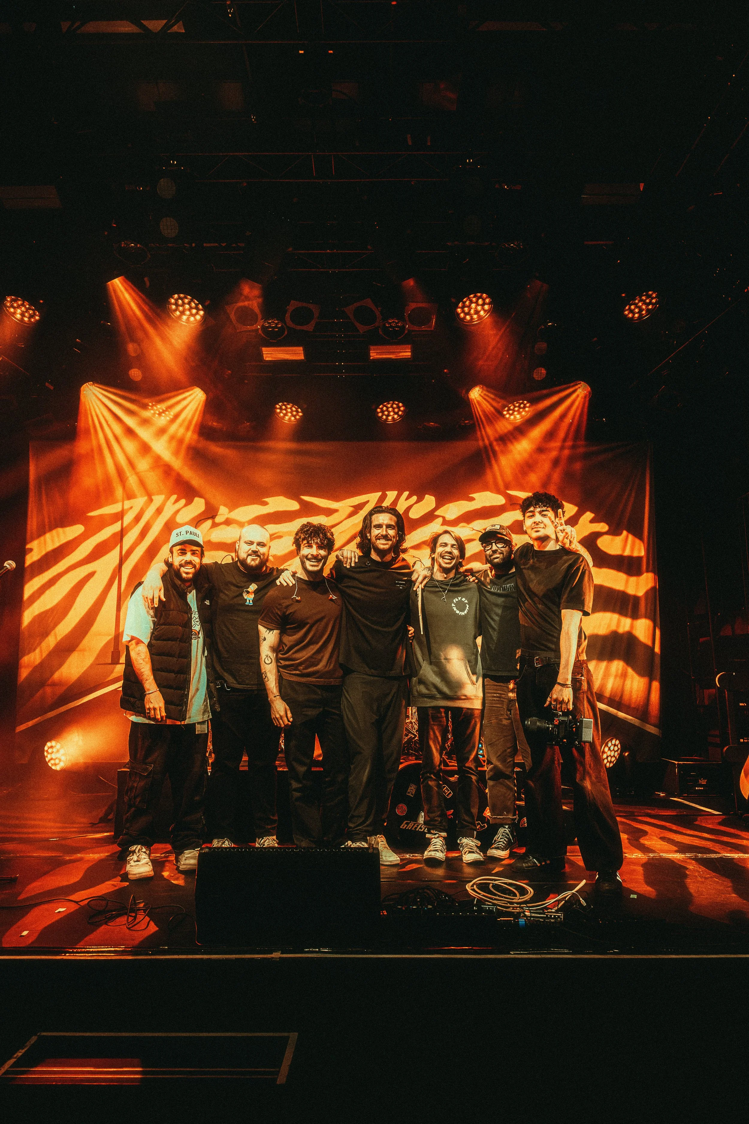 A group of seven men on stage during a concert, standing shoulder to shoulder smiling with arms around each other, with vibrant orange stage lighting and a background featuring a swirling orange pattern.