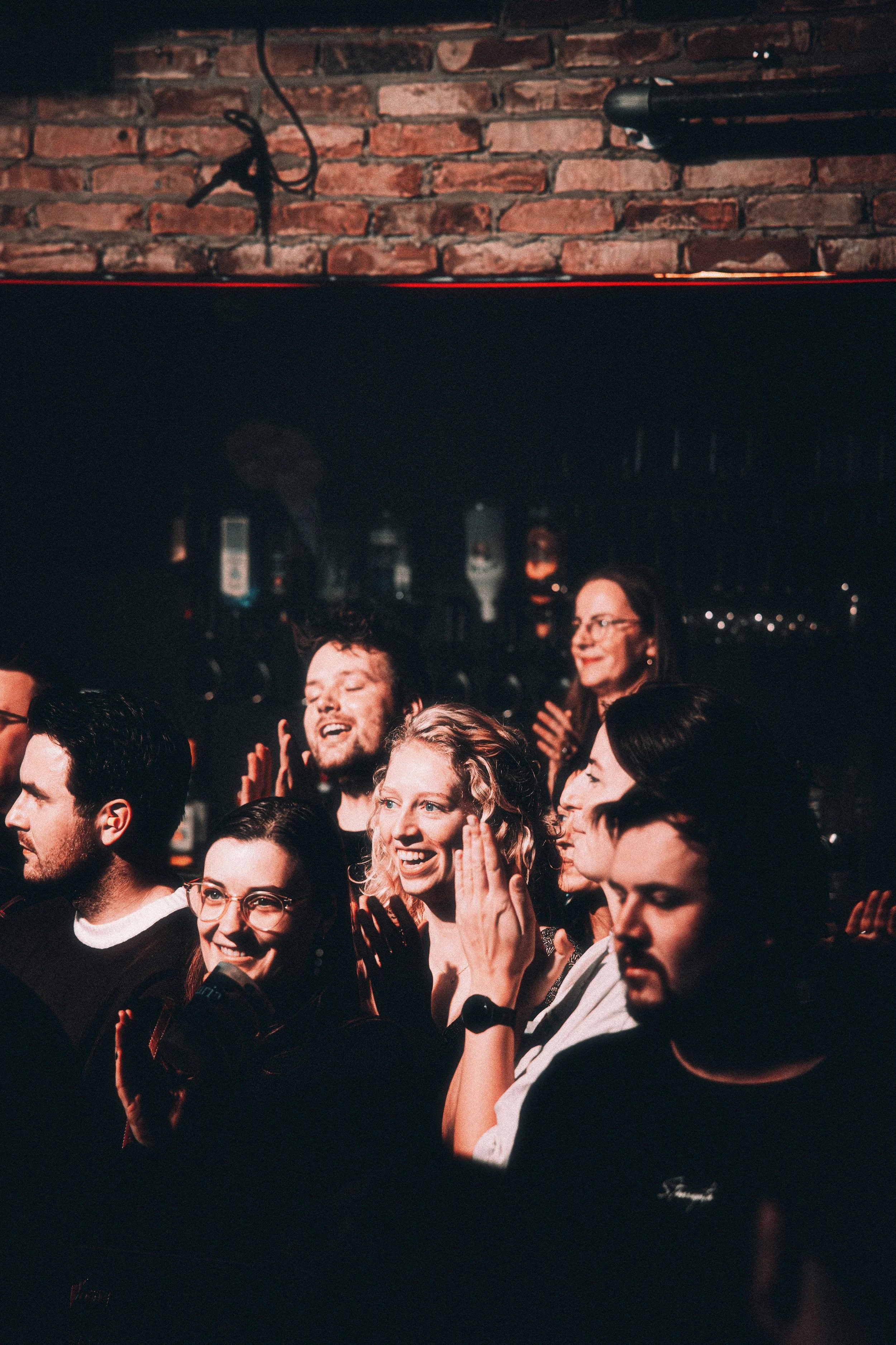 Group of people at a comedy show or concert, clapping and smiling in a dimly lit venue with a brick wall background.