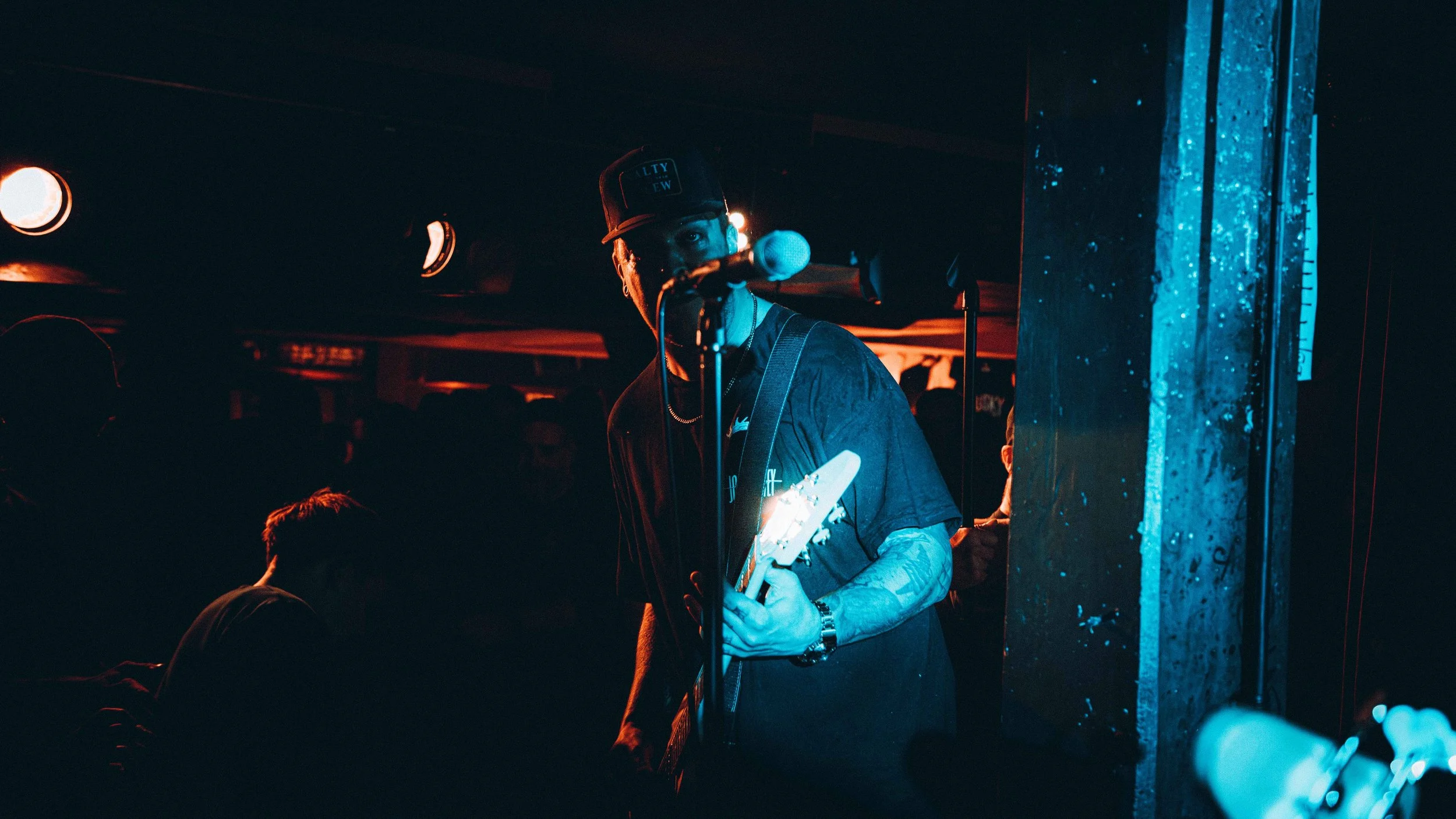 A musician holding a guitar performs on stage, wearing a baseball cap and t-shirt, illuminated by blue and orange stage lights.